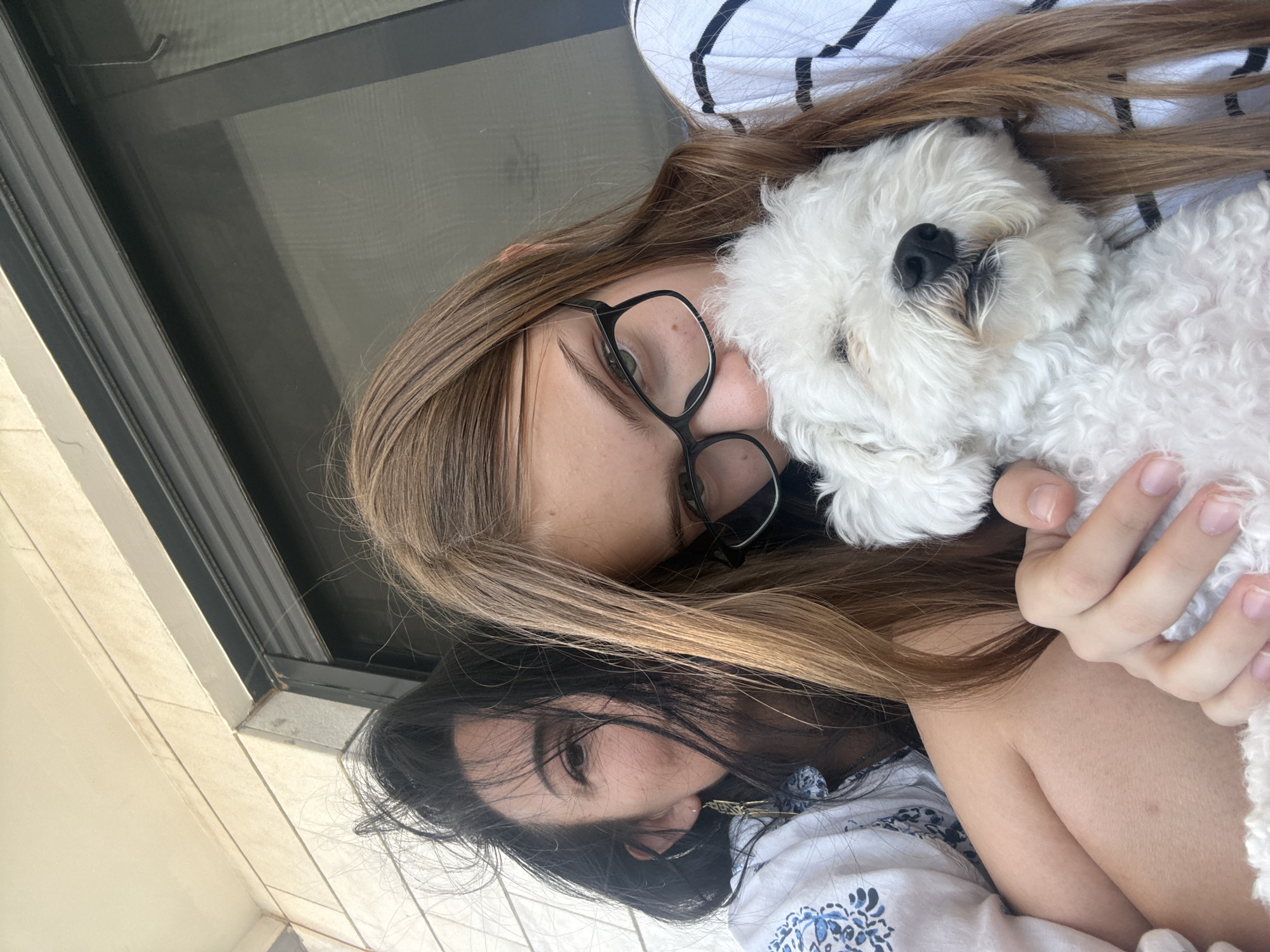 Two females and a small white dog are depicted in this close-up, outdoor selfie. The female in the foreground, identified by her long brown hair and glasses, is holding the dog. The second female, with long dark hair, is partially visible behind the first. The individuals are on what appears to be a balcony or patio, with a textured wall and a window frame visible in the background, suggesting an urban setting. The lighting indicates it is daytime. The overall composition suggests a candid moment shared between the subjects and their pet.