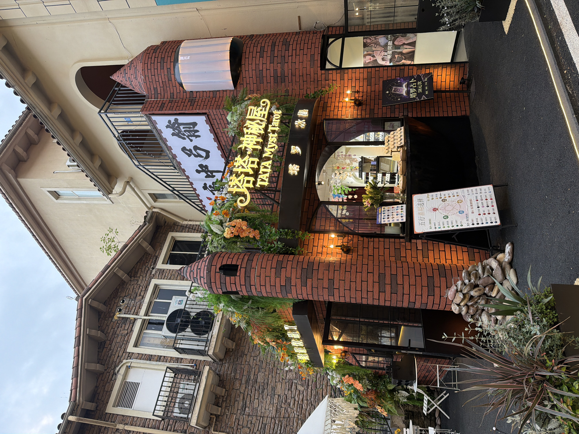 The image shows the facade of "TATA Myst Tarot", a tarot reading establishment located in the Chongqing High-tech Zone, China. The building's architectural style is unusual, with brickwork, rounded towers, and decorative floral arrangements framing the entrance. A large illuminated sign in both Chinese and English identifies the business. Additional signs advertise tarot card reading sessions. The building is multi-storied with a balcony and decorative stone elements. The surroundings include a dark paved area and vegetation, suggesting an urban or semi-urban setting.