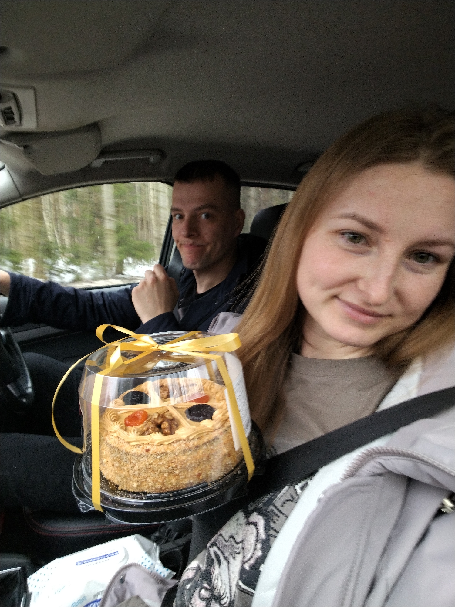 A Caucasian man and woman are sitting in the front seats of a moving car. The man is driving, wearing a dark jacket, and looking at the camera with a slight smirk. The woman is in the passenger seat, wearing a grey jacket and a seatbelt, holding up a cake in a clear plastic container tied with a yellow ribbon. The cake is decorated with nuts, dried fruits, and a light brown cream frosting. Under the cake are wipes. Outside the window, blurred trees are visible. The location is presumably Chaikovsky, Russia.