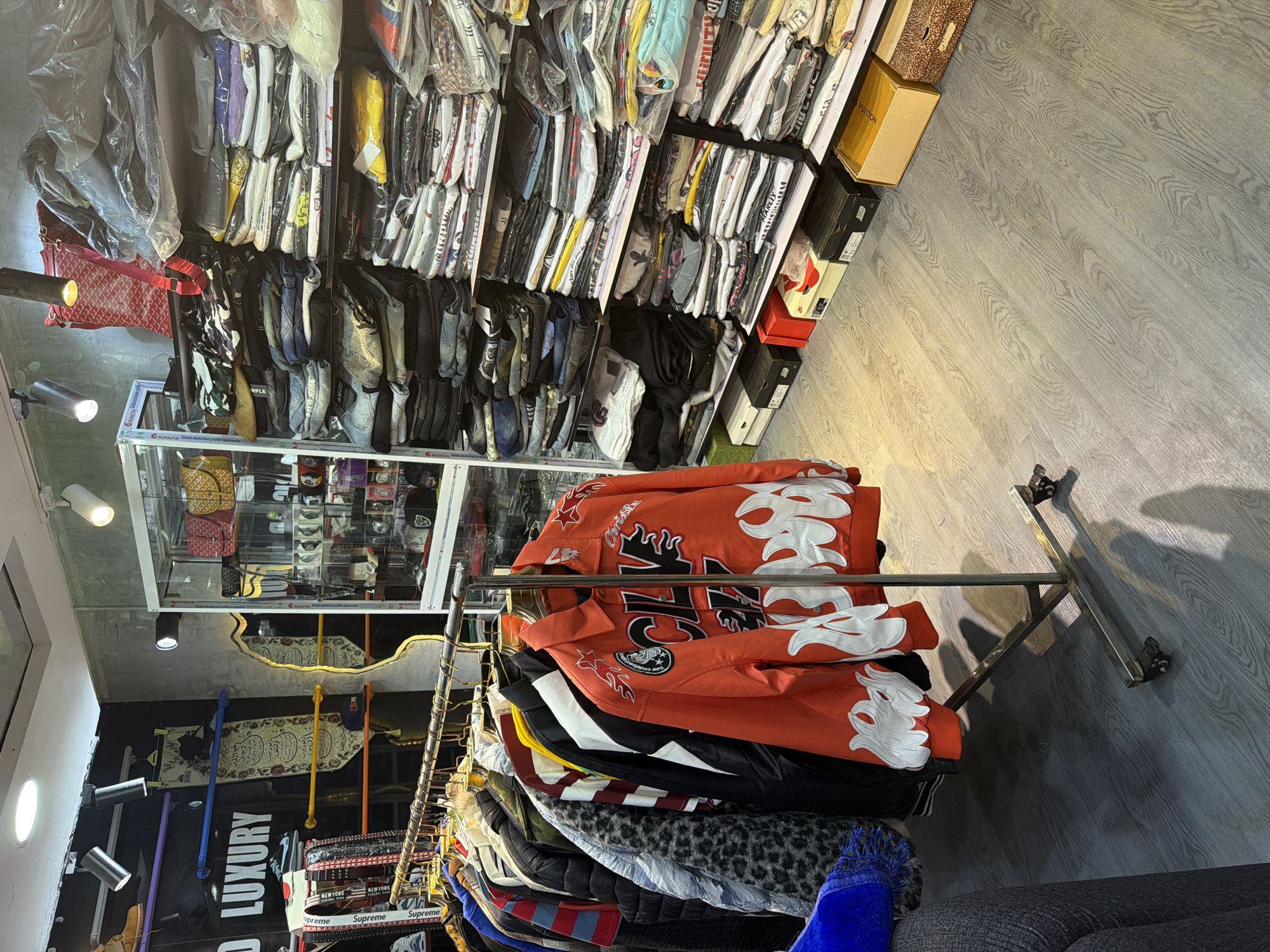 The image depicts an indoor scene, likely a retail establishment, with clothing and accessories displayed on shelves and a clothing rack. The flooring is a grey, wood-grain laminate. To the left, a black wall features the text "D LUXURY" in large white lettering, accompanied by a purple bar and what appear to be decorative textiles or banners hanging from yellow rods. A silver clothing rack stands in the foreground, holding a variety of garments, with a prominent orange jacket with flame-like graphics on the front and sleeves. Behind the rack, a glass display case shows various items, possibly bags and watches. The shelves to the right are filled with neatly folded clothing, some packaged in plastic. Various boxes, including what appears to be a "LOUIS VUITTON" box, are stacked on the floor. The lighting suggests overhead spotlights and accent lighting within the display case. The overall impression is of a fashion boutique.