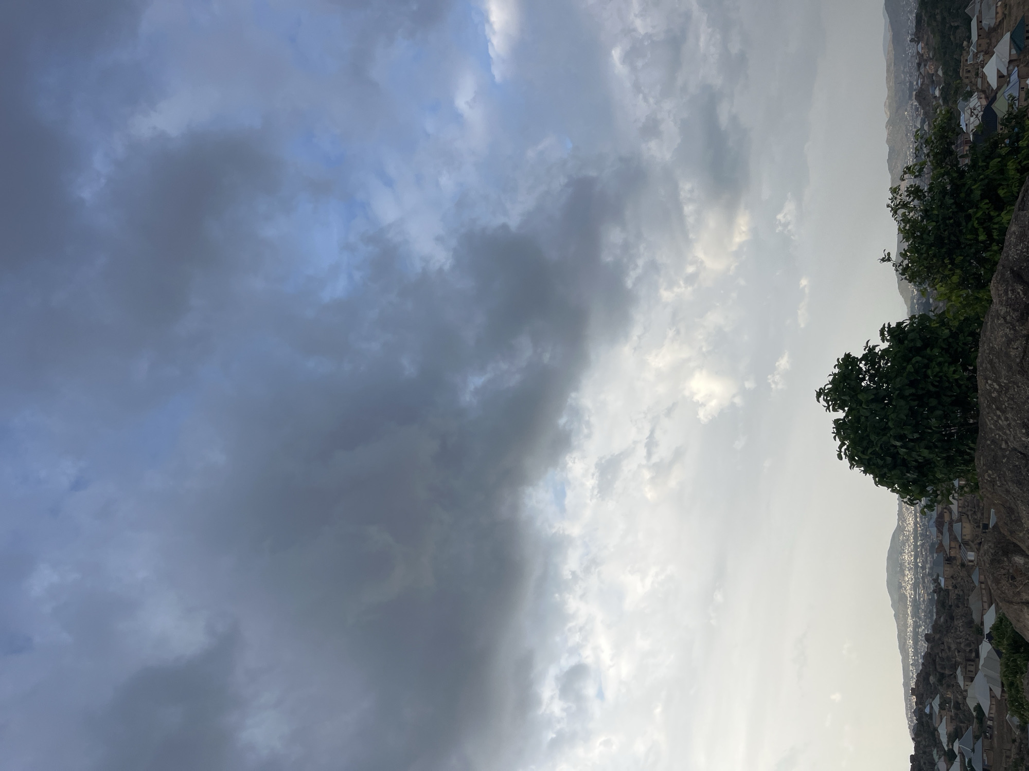The image shows a view from a high point overlooking a part of Jos, Nigeria. The sky is dominated by heavy, grey clouds to the left, contrasted by lighter, brighter clouds toward the right. The foreground features rocks and a tree with dark green foliage. Below the rocks and trees, a densely built urban area with houses and buildings stretches toward the horizon. A low mountain ridge is visible in the distance.