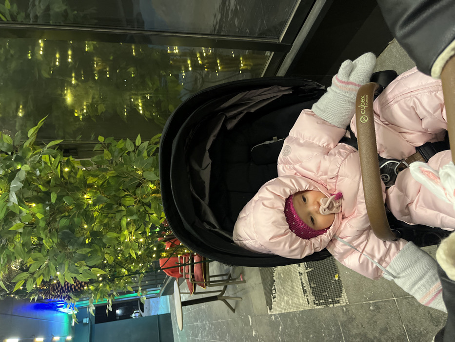 A baby dressed in a pink snowsuit, gray gloves, and a sparkly pink hat lies in a black stroller. The stroller has a Cybex logo on the bar. The baby has a pacifier in their mouth. A bush with yellow fairy lights is beside the stroller. A table and chairs are partially visible behind the stroller. The scene appears to be outdoors at night.