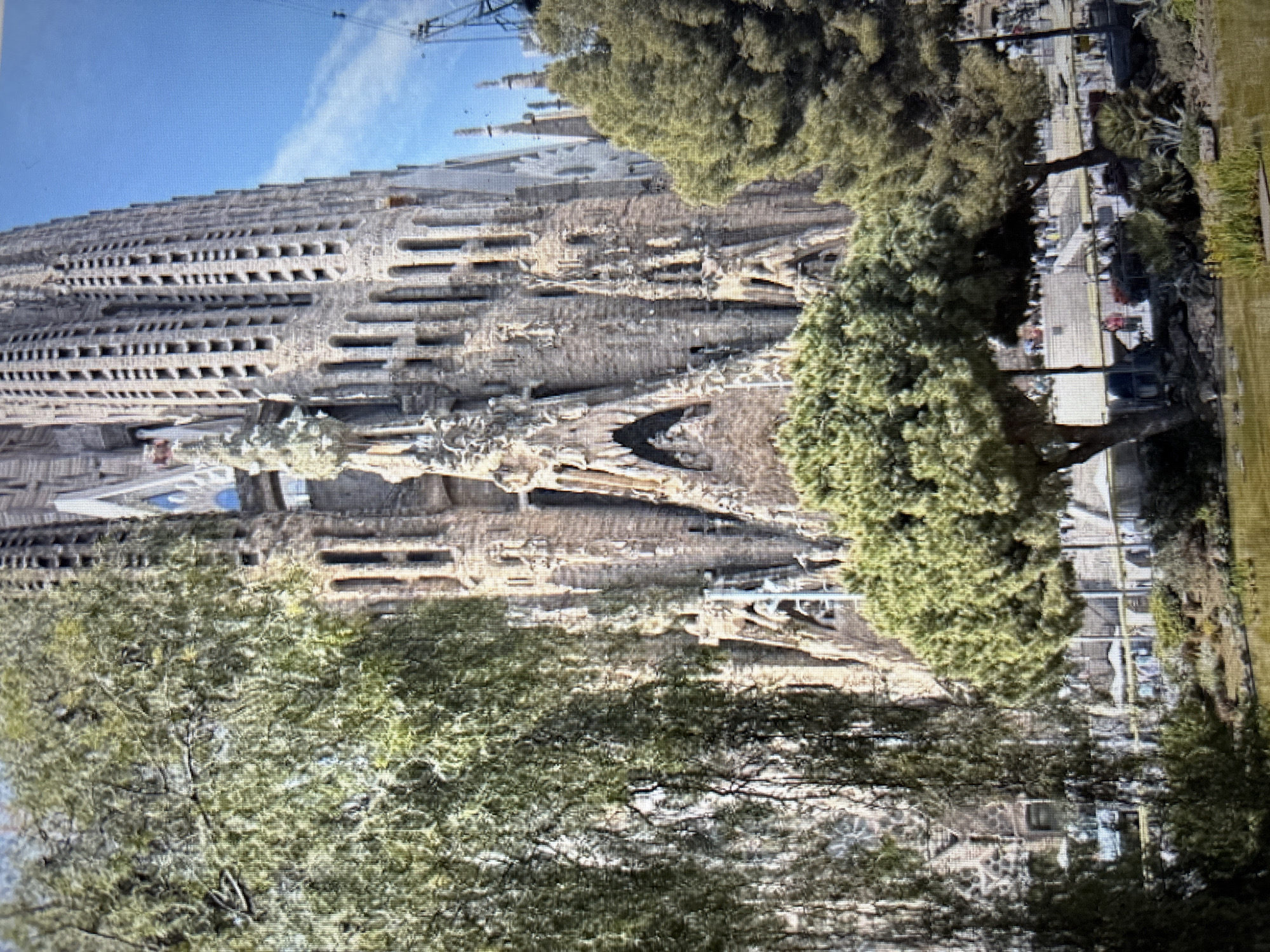 The image depicts the Sagrada Familia basilica, a renowned architectural landmark situated in Barcelona, Spain, not Sofia, Bulgaria. The basilica is a large, intricately designed structure with numerous spires and facades made of stone. In the foreground, there is a body of water, possibly a pond or lake, with some aquatic plants. Lush green trees and foliage surround the water and partially obscure the view of the basilica. The sky above is blue with some wispy clouds. In the background, beyond the trees and basilica, there are hints of human activity, including what appear to be pathways, railings, and scattered individuals, suggesting a public area or park surrounding the monument. The overall scene captures a sunny day with a blend of natural elements and monumental architecture.
