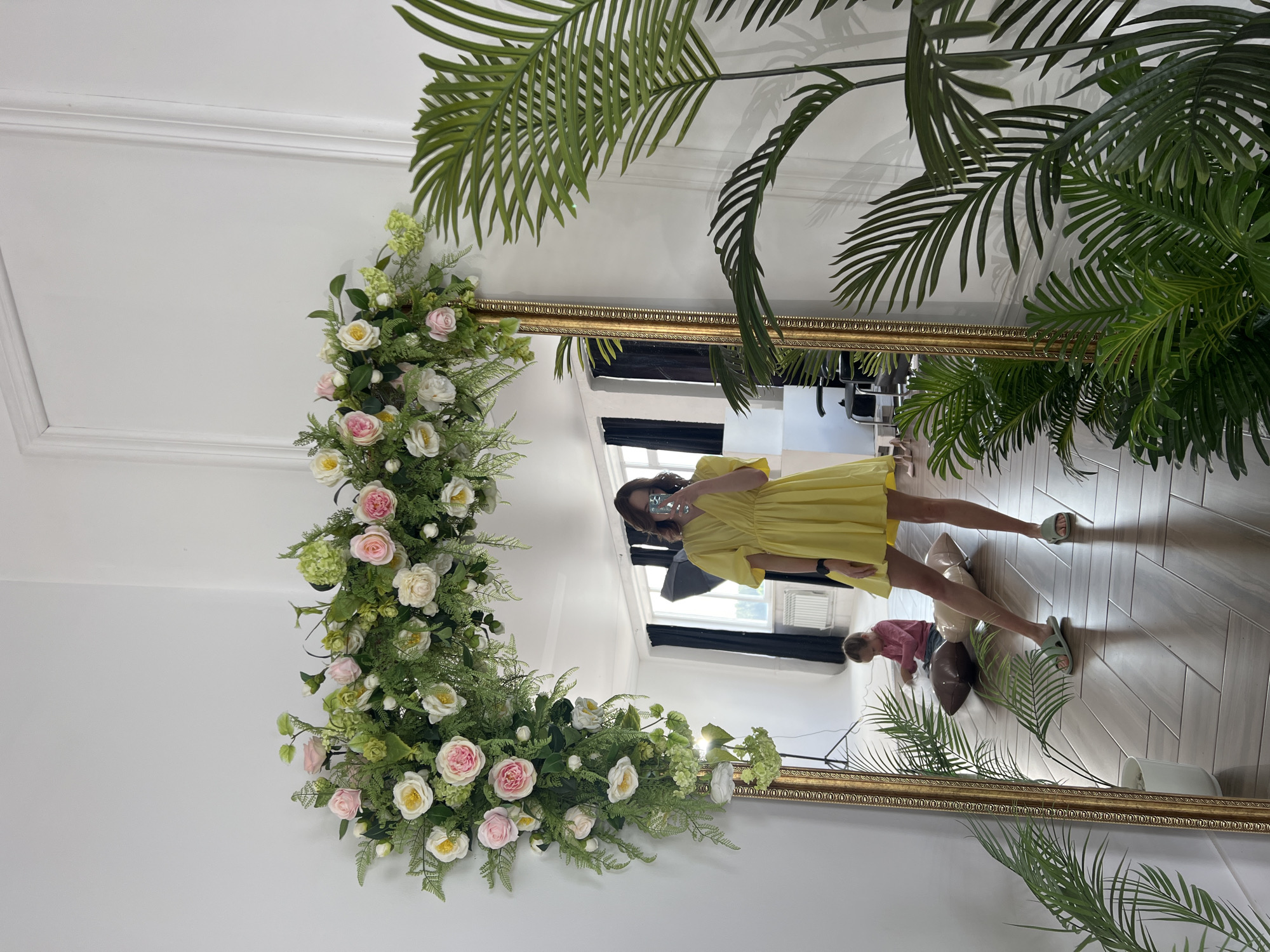 A person in a yellow dress is taking a selfie in front of a large, ornate gold-framed mirror. The mirror is decorated with a garland of white and pink flowers and greenery. A palm plant is positioned to the right of the mirror, partially framing it. The person is holding a phone up to their face, obscuring it. The reflection in the mirror shows a bright room with a window and a person in a pink shirt playing on the floor. The room appears to have light-colored walls and tiled flooring. The location appears to be in Chaikovsky, Russia.