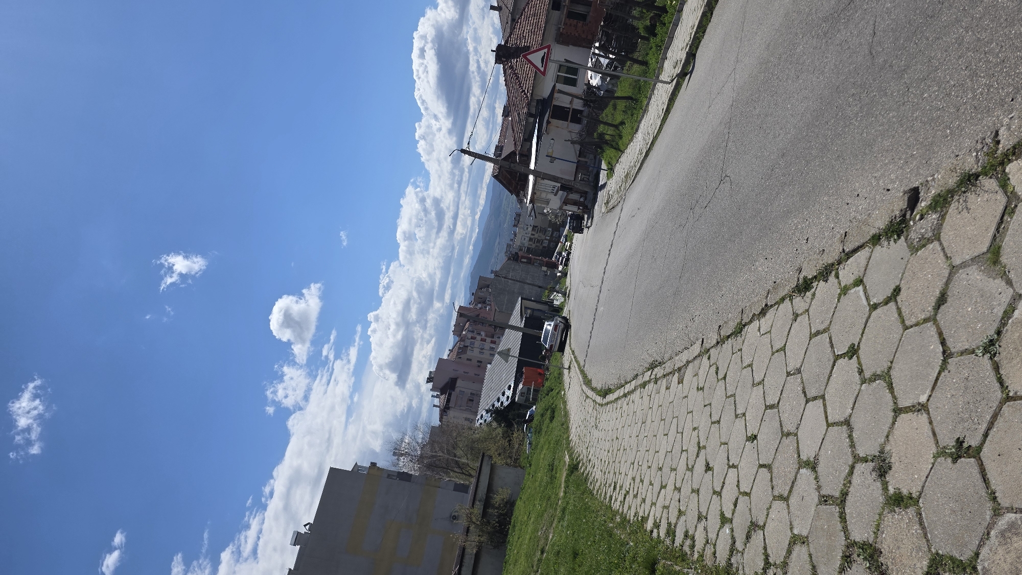 The image shows a street scene in Blagoevgrad, Bulgaria. A gray asphalt road curves slightly, bordered by a hexagonal-patterned concrete sidewalk. Grass and weeds grow in the gaps of the sidewalk. Buildings line the street, including a multi-story apartment building with a yellow facade, residential homes, and a building with a pitched roof marked by a red and white road sign. Several parked cars are visible along the side of the road. In the background, there are more buildings and mountains. The sky is blue with fluffy white clouds.