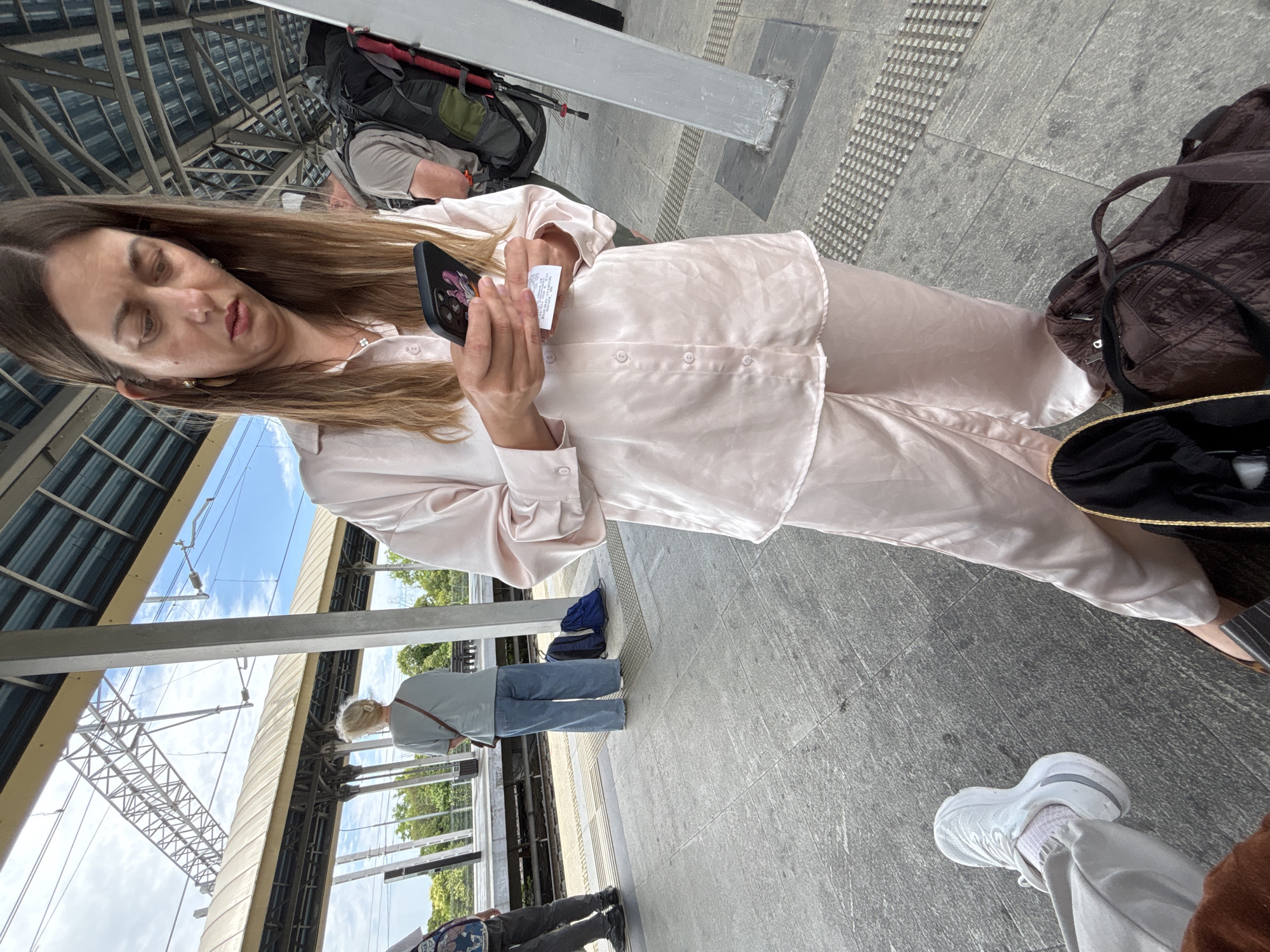 A medium shot captures a woman dressed in a light pink satin pajama set standing on a train platform. She is looking down at her phone, which displays a cartoon character. In her left hand, she holds a receipt. To her right, a person with a large backpack is visible, and further behind them, another individual in a light blue t-shirt and jeans stands near two blue backpacks. The platform is paved with gray tiles, and a covered walkway with a metal roof extends over the platform. The background shows railway tracks and trees. The image does not depict the city of Sirius, Russia.