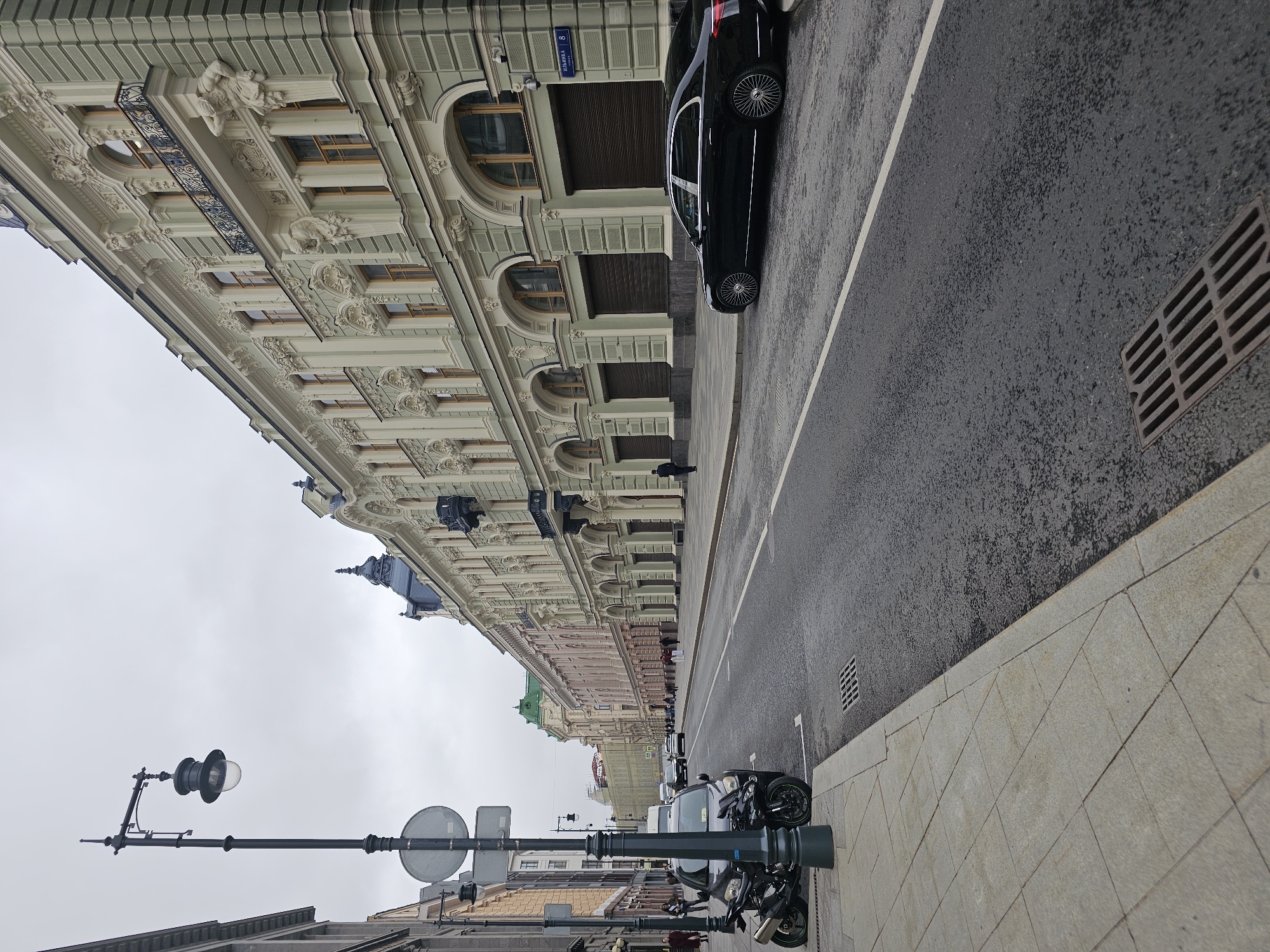 The image shows a street scene in Moscow, Russia, likely in a historic district, given the ornate architecture of the buildings. The overcast sky suggests it is either a cloudy day or early morning.
On the left side of the street, there is a sidewalk paved with large, light-colored square tiles. A black lamppost with a classic design stands prominently on the sidewalk, next to a traffic sign. A black motorcycle is parked on the side of the road, partially on the sidewalk.
The street itself is asphalt, appearing wet, indicating recent rain. A white lane divider line runs down the middle of the road. A black luxury sedan is parked on the right side of the road.
The buildings lining the right side of the street are tall and ornate, with a light green facade and intricate detailing, including columns, arches, balconies with wrought-iron railings, and sculptures. Several windows are visible, many with arched tops. Some ground-level entrances appear to be garage doors. A street sign with Cyrillic lettering and the number "8" is mounted on the building.
Further down the street, other buildings are visible, some with different colored facades and green roofs. A few pedestrians are walking on the sidewalks and crossing the street in the distance. Several cars and a white van are driving on the street. The overall impression is of a well-maintained, upscale urban environment.