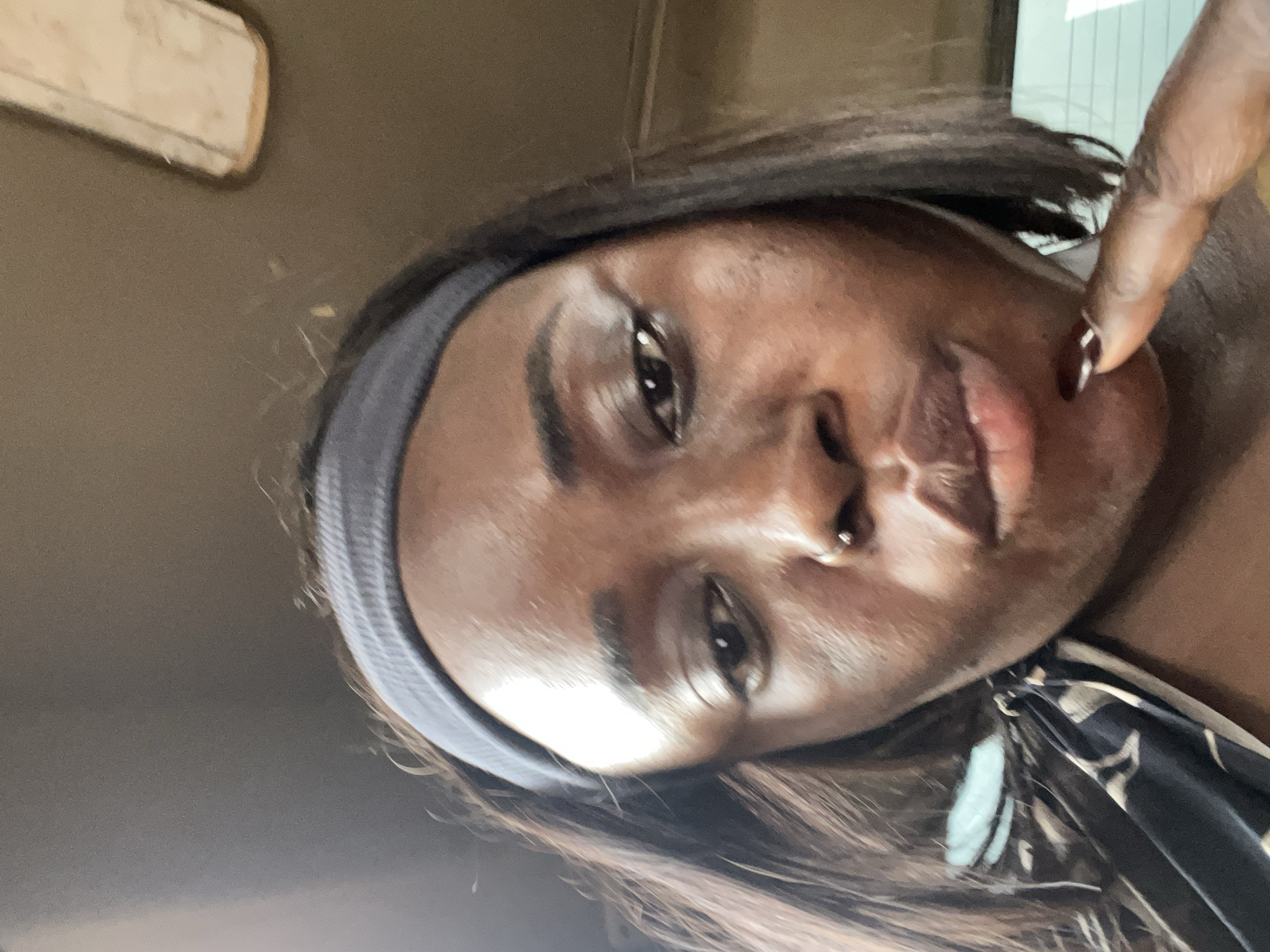 A close-up, rotated, portrait-style selfie of a Black woman. She is wearing a grey headband and a black and white patterned garment. Her skin is smooth and appears to have some makeup on. Her eyes are dark and her lips are full. She has a nose ring. One of her fingers is touching her cheek near her nose. The background is out of focus and appears to be indoors, possibly a car. The lighting suggests it could be daytime. There is no visible text.