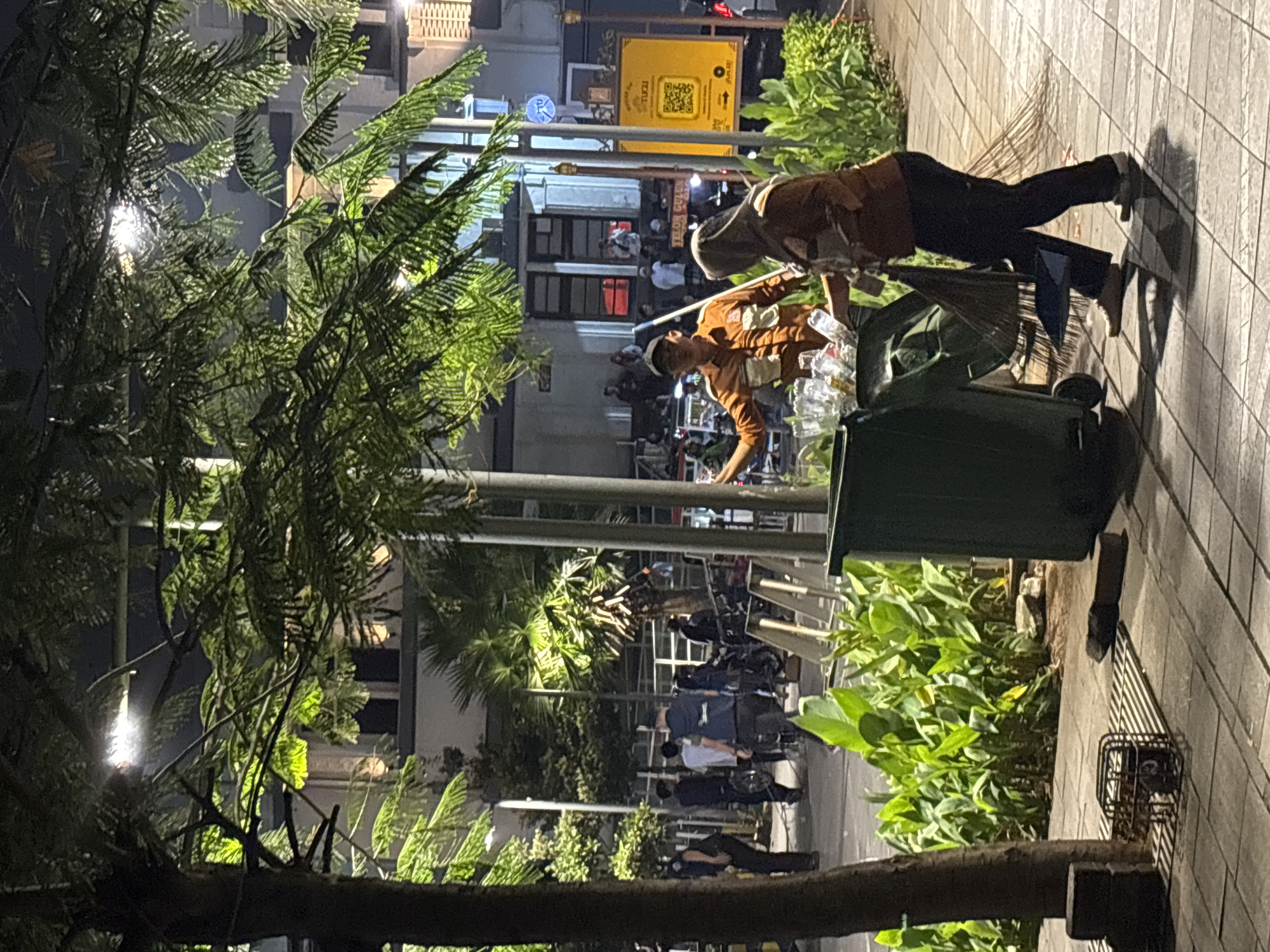 Two individuals, a man and a woman, are working near a large green trash bin on a tiled sidewalk in West Jakarta, Indonesia. The man, wearing an orange uniform, is reaching into the bin, appearing to sort or retrieve items. The woman, also in an orange uniform and a headscarf, is holding a broom and dustpan, seemingly preparing to sweep or clean the area. The scene is illuminated by overhead streetlights and lights from buildings in the background. In the background, several other people are visible, some standing and others near motorcycles, suggesting a public area or thoroughfare. Lush green plants border the sidewalk. The time appears to be evening or night.