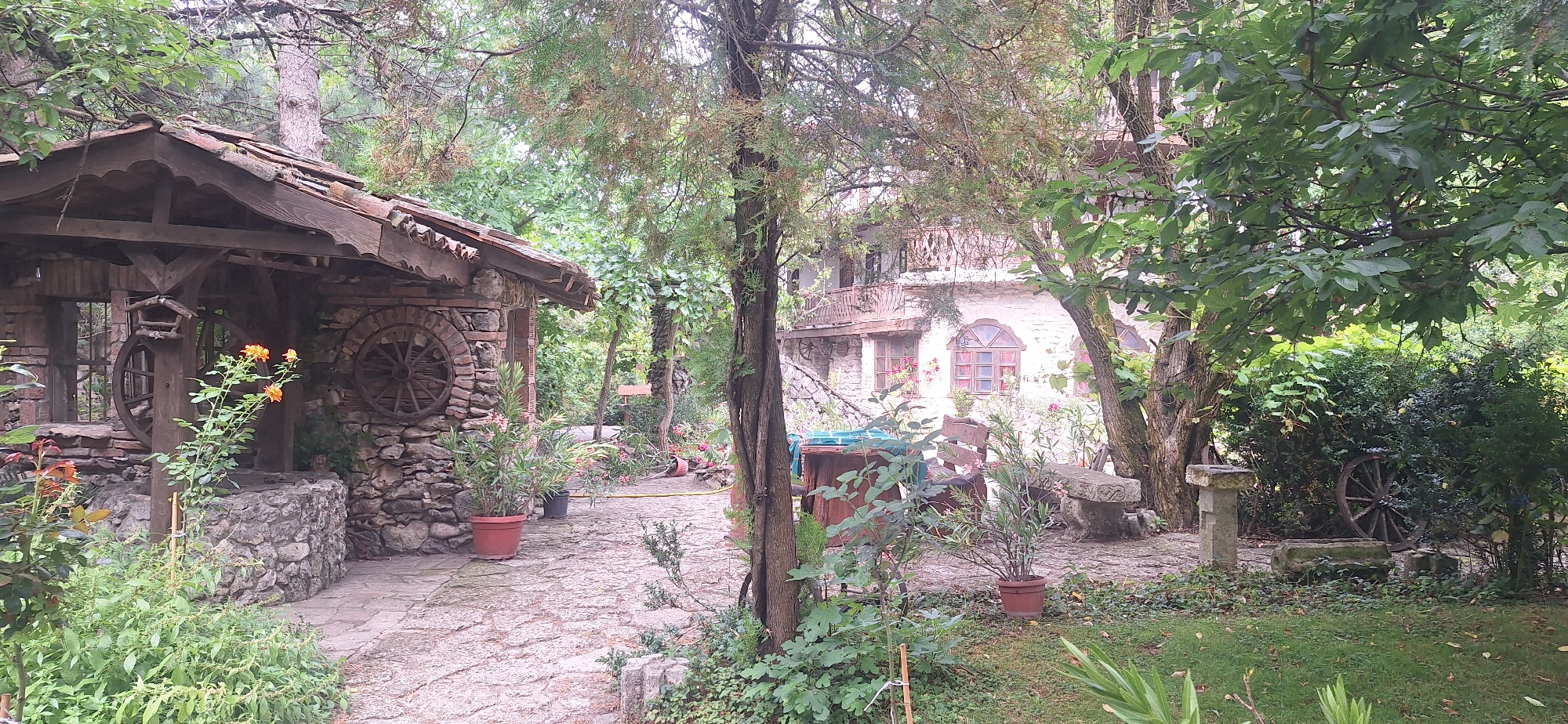 The image depicts a rustic, wooded outdoor area with two stone and wood structures. To the left, a small, open-sided building with a tiled roof and stone walls features decorative wooden wagon wheels. A well with a wooden frame and a bird feeder is situated in front of it. Orange flowers bloom nearby. To the right, a larger, multi-story building constructed with white stone has wooden balconies and arched windows with red accents. The area between the structures is paved with stone and surrounded by lush greenery, including trees, bushes, and various plants. A wooden bench, a stone carved pillar, and more wagon wheels are scattered throughout the grounds. The overall scene suggests a quaint, possibly historical, rural setting. The location is identified as Kalimantsi, Bulgaria. There are no people visible in the image.