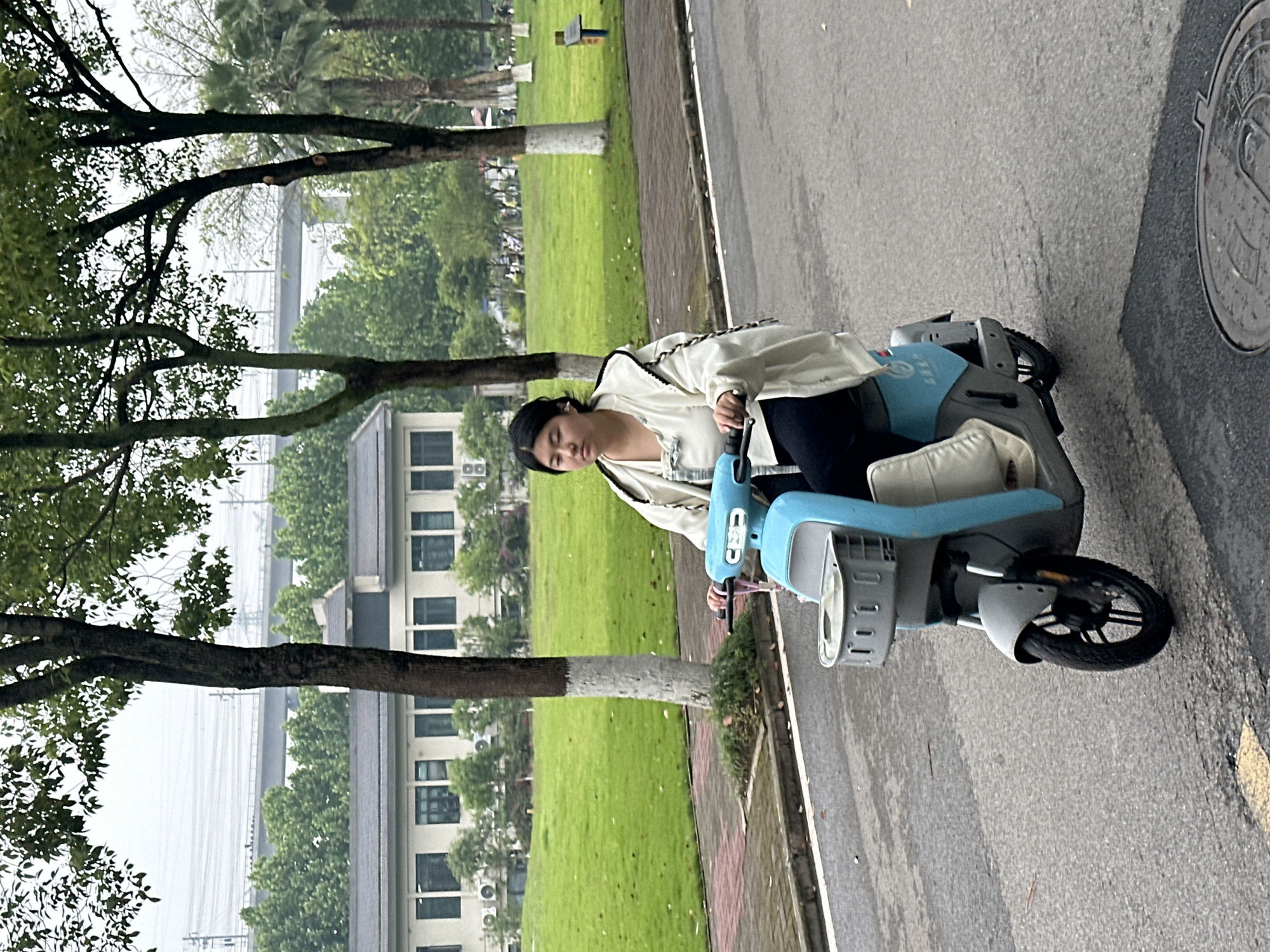 A young woman is depicted seated on a light blue and grey electric scooter on what appears to be a damp paved surface, likely a road or wide pathway. She wears a light-colored jacket over a white top and dark trousers, with her hands on the scooter's handlebars. Her dark hair is pulled back, and she looks downwards with a neutral expression.
To the woman's left, a green grassy area is visible, dotted with trees, some of which have their lower trunks painted white. Behind the trees and grass are multi-story buildings with numerous windows, and in the far background, a hazy structure, possibly a bridge or distant urban infrastructure, can be seen beneath an overcast sky. Palm trees are also present in the background.
To the woman's right, the asphalt road continues, featuring a circular metal manhole cover embossed with intricate Chinese characters. Faint white Chinese text, possibly "哈啰" (Hello), is visible on the blue body of the scooter, suggesting it might be a shared mobility vehicle. The overall setting appears to be an outdoor urban or campus environment in Jiangjin, China, during daytime with overcast weather. The ground's damp appearance suggests recent rainfall.