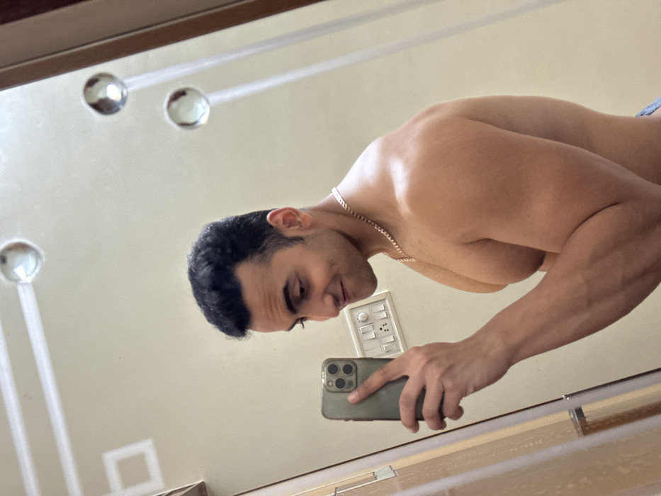 A man takes a mirror selfie. He has dark hair, tan skin, and a gold chain. He is shirtless, showcasing muscular arms and shoulders. The phone in his hand is a green iPhone. Behind him on the wall, there's a white electrical panel, and the mirror reflects a decorative design of silver spheres. The perspective is slightly tilted, giving a low-angle view.