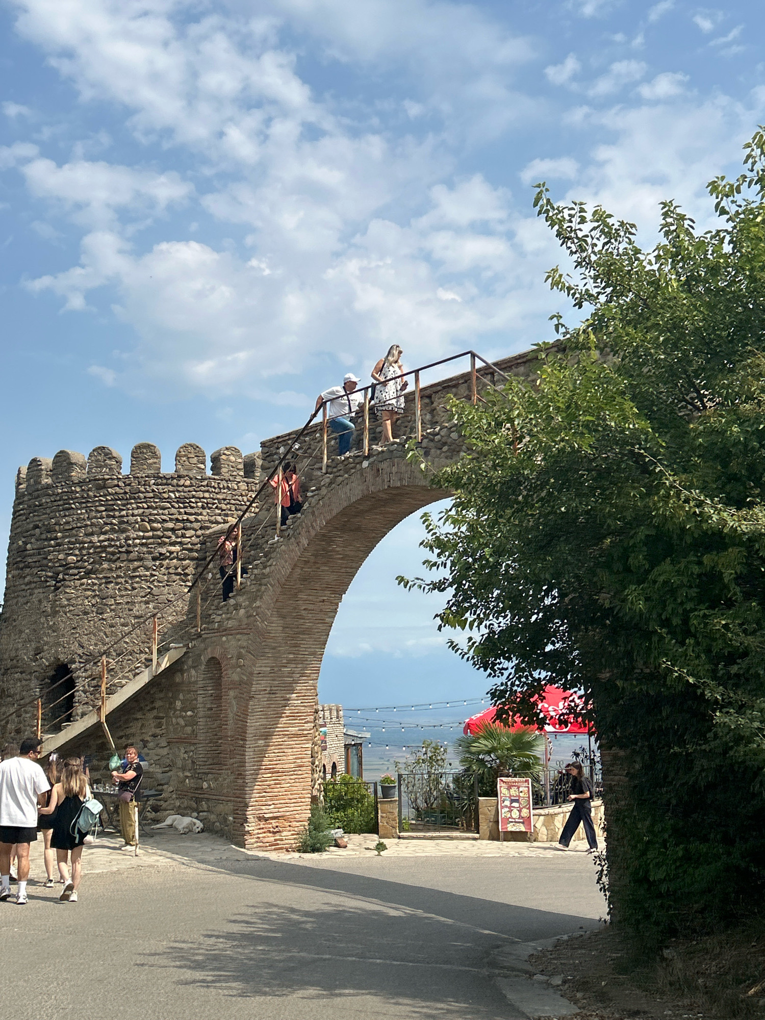 A tour group walks across a brick archway and stone tower, overlooking a distant landscape. The tower is made of rough-hewn stone with crenellations at the top, and a staircase ascends to the archway. Several people are on the stairs and walkway. On the ground level, a white dog sleeps near a stone wall. To the right of the archway, a red umbrella shades an outdoor dining area. A woman in dark clothing walks past a restaurant sign. The sky is blue with scattered white clouds. The location is the city of Sighnaghi, Georgia.