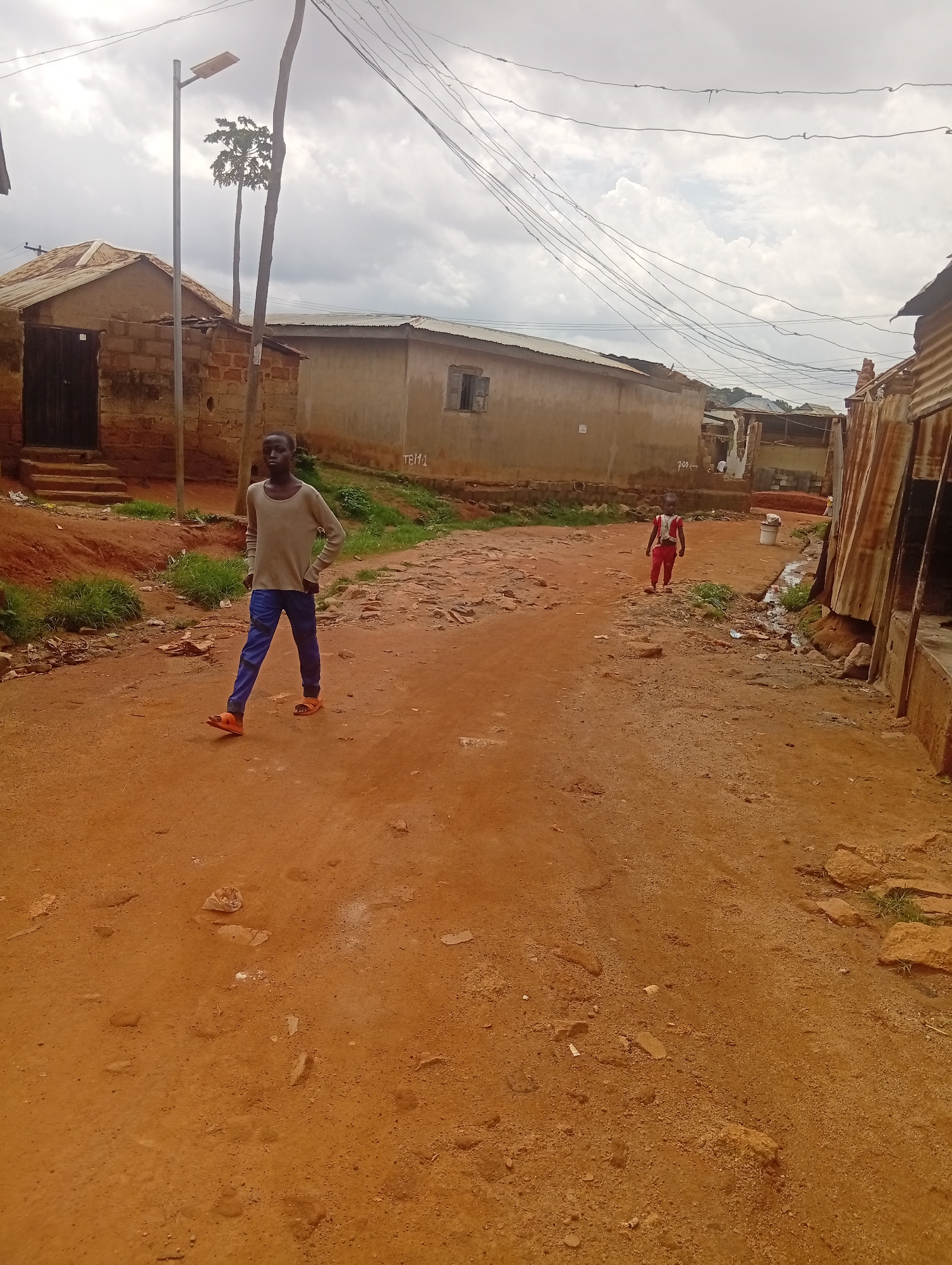 A young boy in a light-colored sweater and blue pants walks across a dirt road in a Nigerian village. He wears orange sandals and has his hands on his hips. Further down the road, another child in a red shirt and pants walks away from the camera. The scene is set in a village with simple, earth-toned buildings, some constructed of brick and others with corrugated metal roofs. A street lamp stands to the left of the frame, and a tangled mess of electrical wires is strung overhead. The ground is a dusty orange, with scattered rocks and patches of green grass. The sky is overcast, suggesting a cloudy day. The atmosphere appears casual, with the children seemingly going about their daily activities in their community. The buildings and unpaved road suggest a rural or semi-urban setting. There is a small stream or drainage ditch on the right side of the road, with some debris visible. The overall impression is of a typical day in a Nigerian village.
