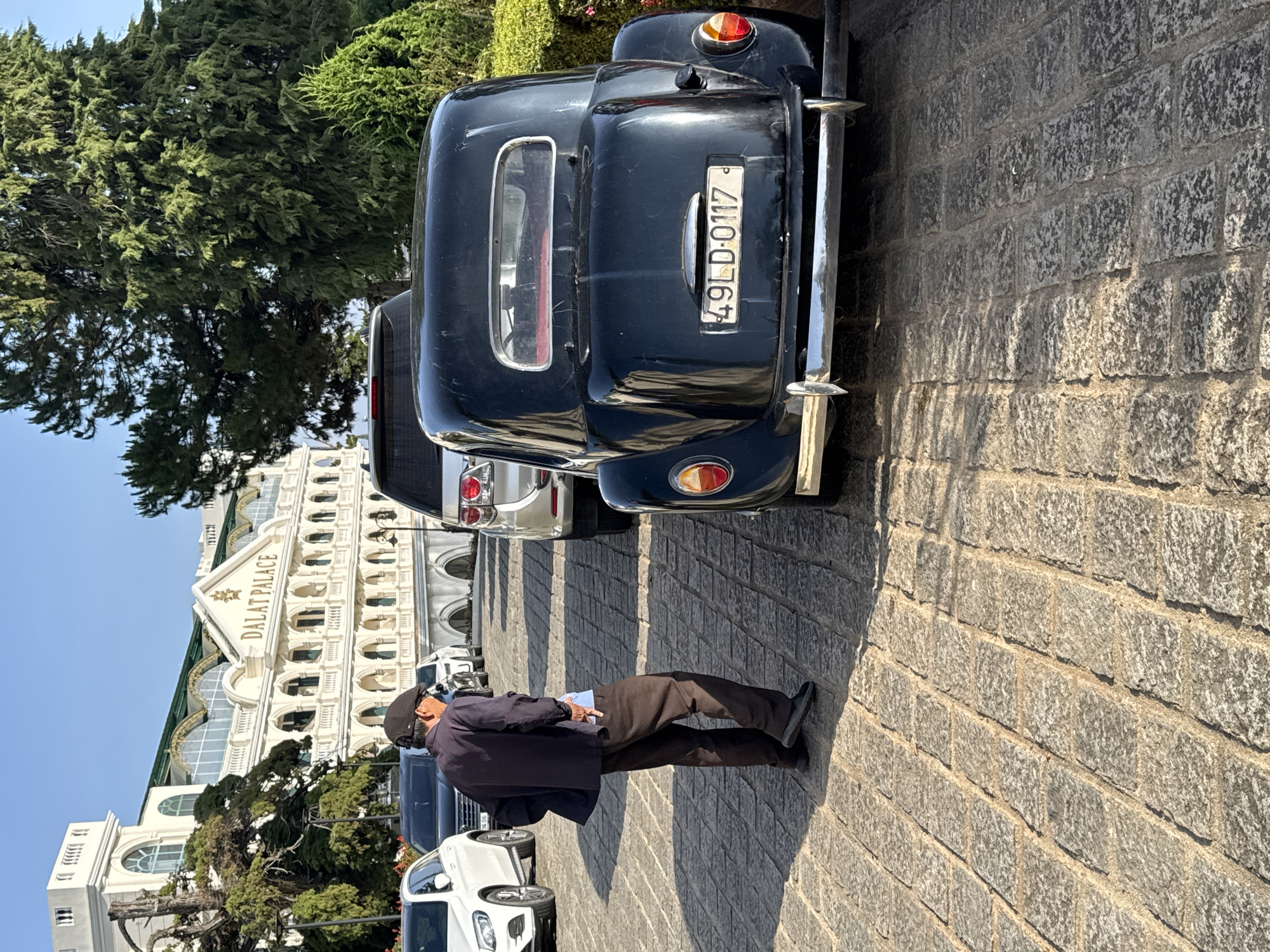 The image shows a man standing near a vintage black car parked in front of the Dalat Palace Hotel in Da Lat, Vietnam. The car's license plate reads "49LD-0117." The man is wearing dark clothing and a hat, holding what appears to be a paper in his hand. The street is paved with square granite blocks. The Dalat Palace Hotel is visible in the background, showcasing its distinctive white facade with multiple archways. The weather appears sunny and clear.