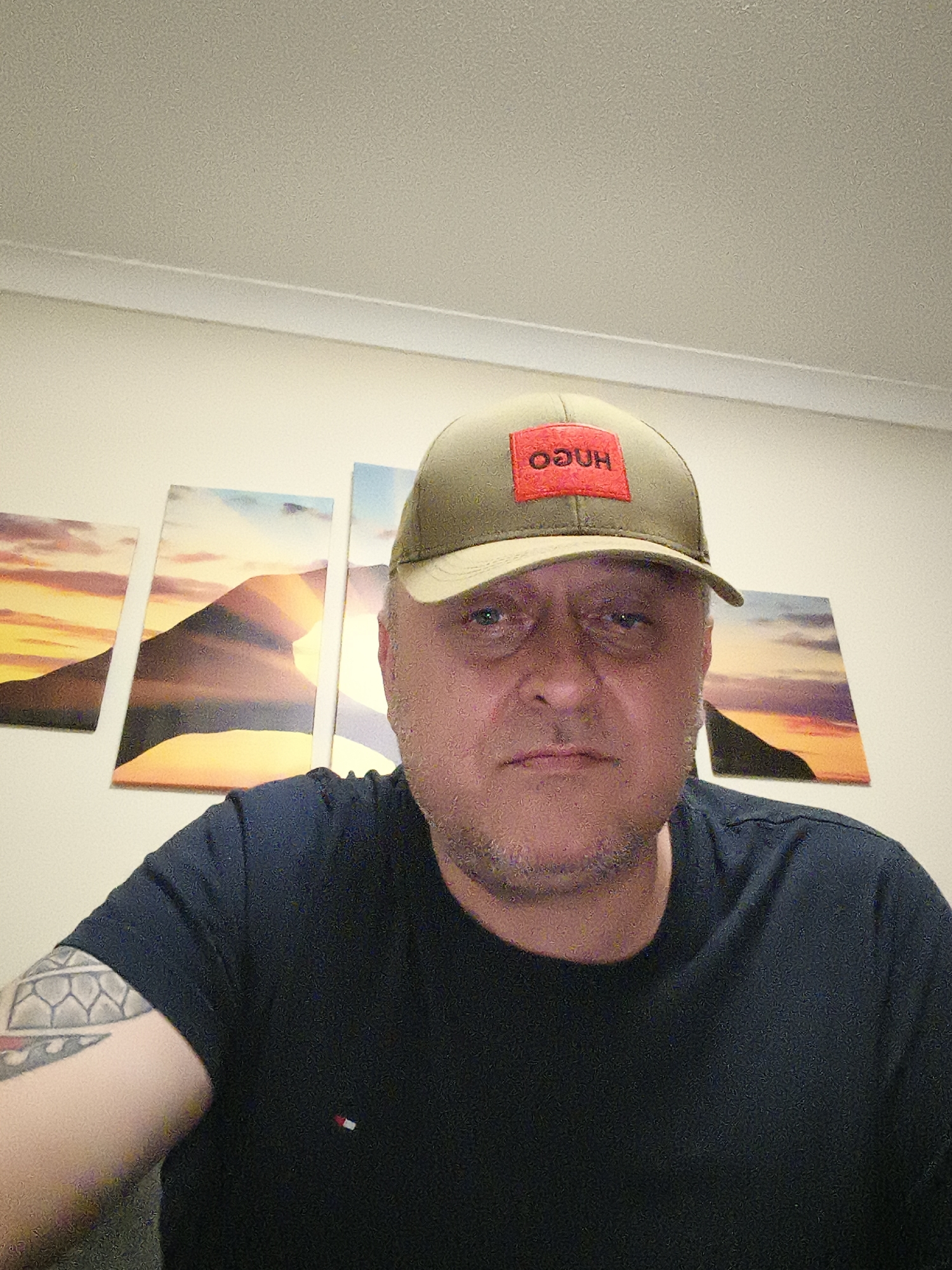 A man, appearing to be middle-aged, is captured in a close-up portrait indoors. He wears an olive green baseball cap with a prominent red patch on the front, displaying the mirrored and inverted text "ODUH" (intended to be "HUGO"). His dark blue short-sleeved t-shirt features a small red, white, and blue flag logo, likely Tommy Hilfiger, on the left chest. A distinctive patterned tattoo is visible on his left upper arm. The man has stubble and maintains a neutral to slightly serious expression as he looks directly into the camera.
Behind him, on a light-colored wall, are four rectangular canvas prints or panels, depicting a cohesive desert or mountainous landscape scene under a sunset or sunrise sky. The ceiling above is also light-colored with white crown molding. The indoor setting, likely a home, is illuminated by artificial light, and the image exhibits some digital noise, particularly in the ceiling and darker areas. This scene is contextualized as taking place in Huntingdonshire, United Kingdom.