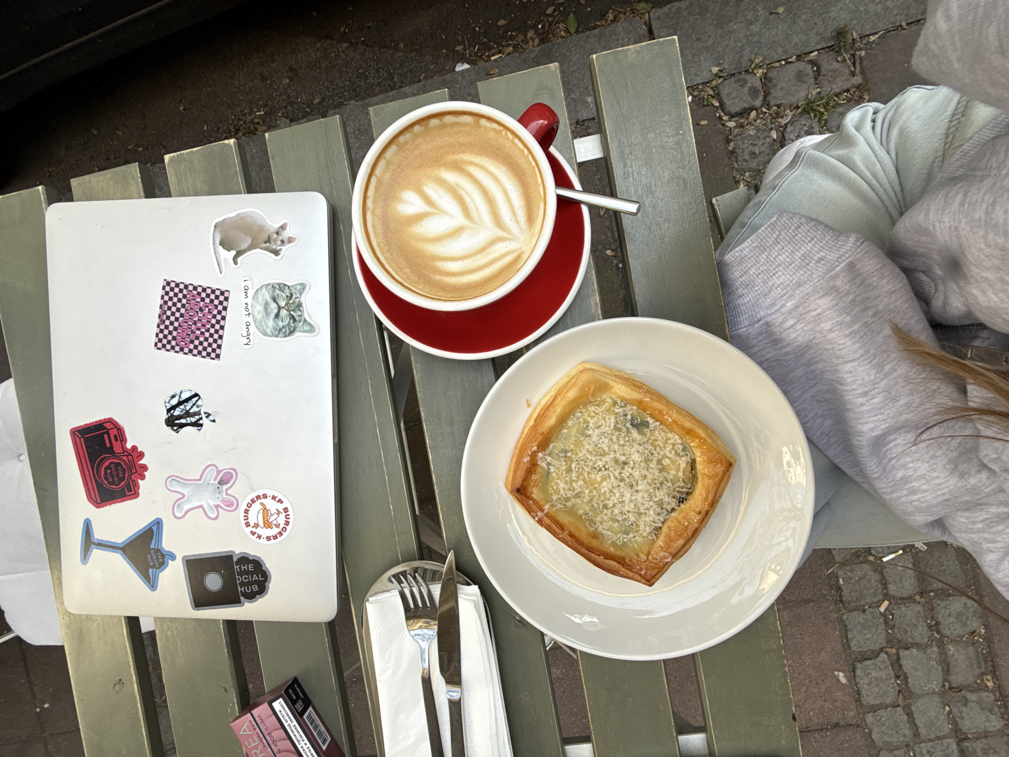 Here's a detailed description of the image:
**General Description:** An overhead shot captures a casual outdoor dining scene. A latte, a savory pastry, and a laptop with various stickers are arranged on a slatted, gray-green table. A pack of IQOS Iluma tobacco sticks is partially visible.
**Objects:**
* **Latte:** A red cup and saucer hold a latte with latte art in the shape of a leaf. A small spoon rests on the saucer.
* **Pastry:** A square savory pastry with visible filling and cheese topping sits on a white plate, accompanied by a knife and fork.
* **Laptop:** The laptop is white with stickers, including cat figures, a camera graphic, a martini glass, and logos for "The Social Hub" and "Burgers-KP".
* **Cigarettes:** A pack of IQOS Iluma tobacco sticks are barely visible.
**People:**
* Clothing is visible. A light gray or blue hooded sweatshirt is partially visible.
**Environment:**
* The table is made of slats. The background includes paved ground and light-colored clothing.
**Location:** While the image itself does not explicitly confirm that the location is Belgrade, Serbia, the presence of an IQOS Iluma cigarette pack with Cyrillic text indicates that the location is likely Serbia. The specific city within Serbia is not explicitly mentioned.