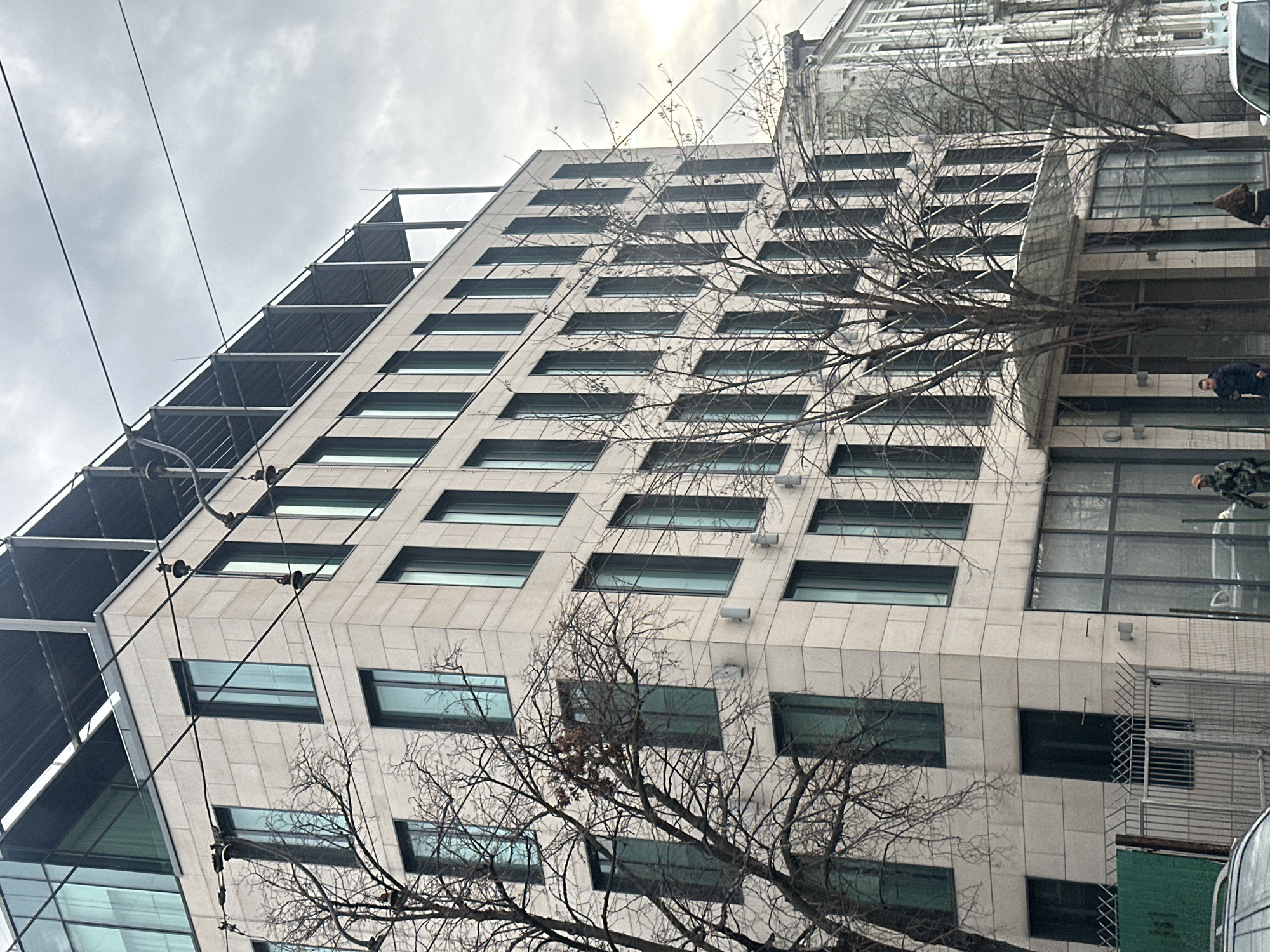 The image shows a multi-story building in Kyiv, Ukraine. The building is constructed of light-colored stone or concrete with rows of rectangular windows. A bare tree stands in front of the building, its branches partially obscuring the view. On the left side of the building, there is a metal structure attached, possibly for scaffolding or architectural detail. Power lines stretch across the upper portion of the frame. On the right side of the building, there are some people in the windows. The sky is overcast, suggesting a cloudy or overcast day.