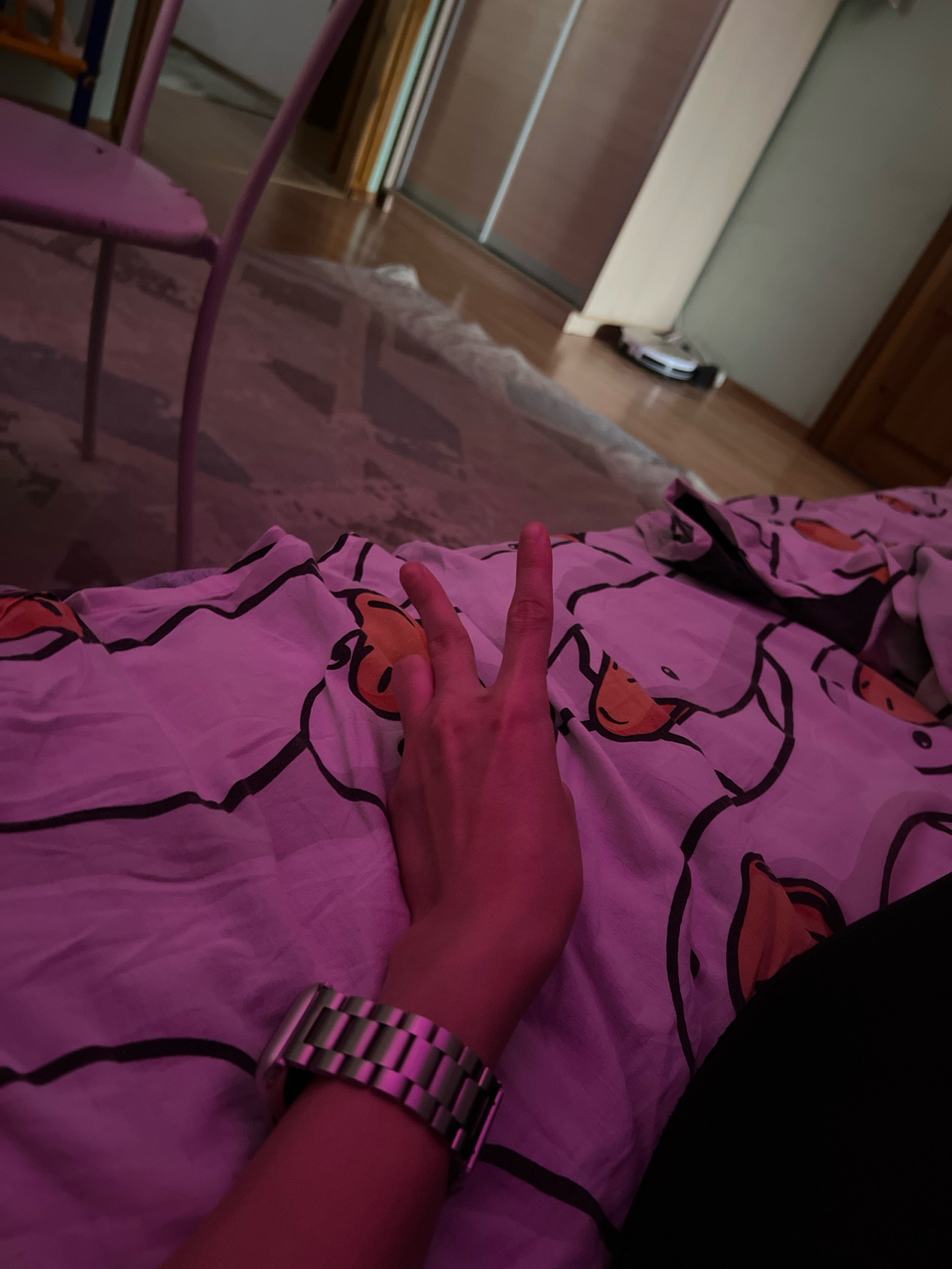 A first-person perspective shot shows a person's left hand with a silver watch on their wrist, resting on a bedspread featuring a white pattern of cartoon ducks. The hand is raised, with the index and middle fingers extended in a peace sign. The bedspread is illuminated with pink light, creating a vibrant and somewhat surreal atmosphere. In the background, a partially visible pink chair and wooden floor are visible, along with a robotic vacuum cleaner near a doorway. The scene is indoors, likely a bedroom, and the overall impression is casual and intimate. The location is identified as Morozovsk, Russia, though this is not visually evident from the image itself.