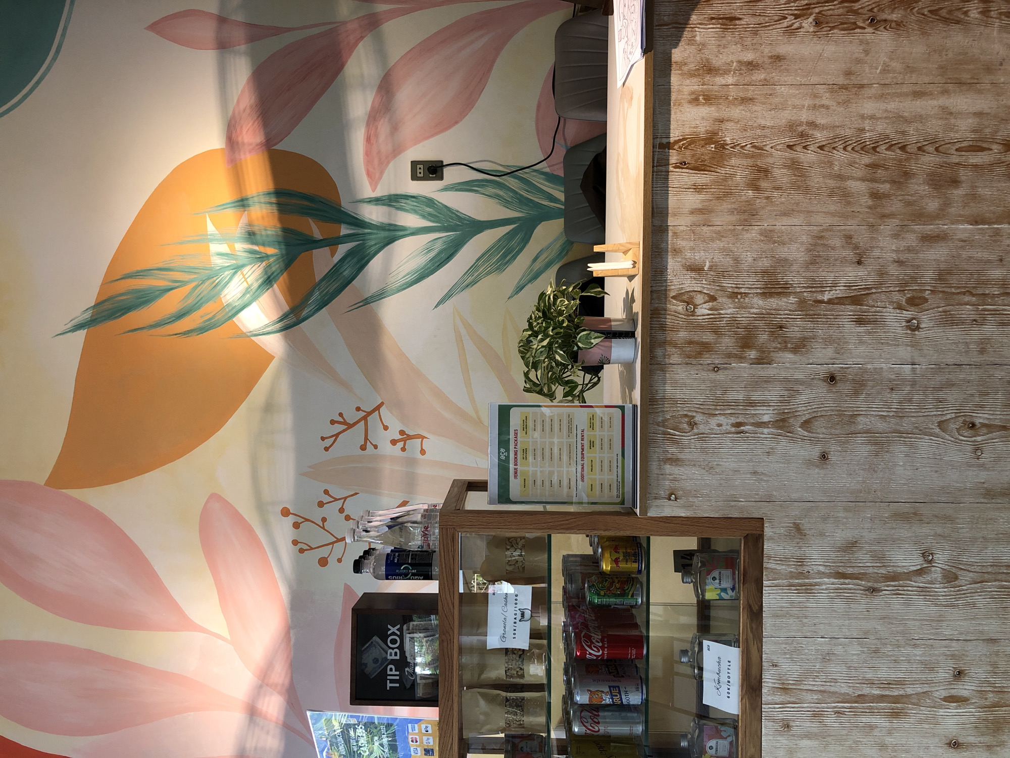 The image shows a counter area in a cafe in Đà Nẵng, Vietnam. A light wood panel wall forms the base of the counter, topped with a light-colored countertop. Behind the counter, a colorful mural of abstract plant shapes in shades of pink, orange, and green covers the wall. On the counter, a small potted plant with variegated green leaves sits in a pink and white pot. To the right of the plant, a wooden napkin holder with two white napkins is visible. A stack of papers is on the right side of the counter, partially obscured.
On the left side of the counter, a glass display case showcases various items. The top shelf of the display case holds a dark wood "TIP BOX" with some cash inside, and several bottles of water. Below this, bags of granola or similar dry goods are displayed, with a handwritten sign that reads "Granola/Cashew 50K/BAG/100G". The bottom shelf of the display case contains bottles of what appears to be Kombucha, with a sign indicating "60K/BOTTLE". Cans of soda, including Coca-Cola and Red Bull, are lined up on the counter in front of the display case.
In the background, there are three grey chairs arranged behind the counter, suggesting a seating area for staff or customers. A black electrical outlet with a cord plugged into it is mounted on the muraled wall. A sign on a stand to the right of the display case lists "VENUE BOOKING PACKAGES" and "ADDITIONAL EQUIPMENT RENTAL" with prices in Vietnamese Dong (VND). Another sign on the left, partially visible, announces "WE ARE GOING CASHLESS 1ST NOVEMBER 2023" and displays logos for various payment methods. The overall impression is a modern, somewhat bohemian cafe interior.