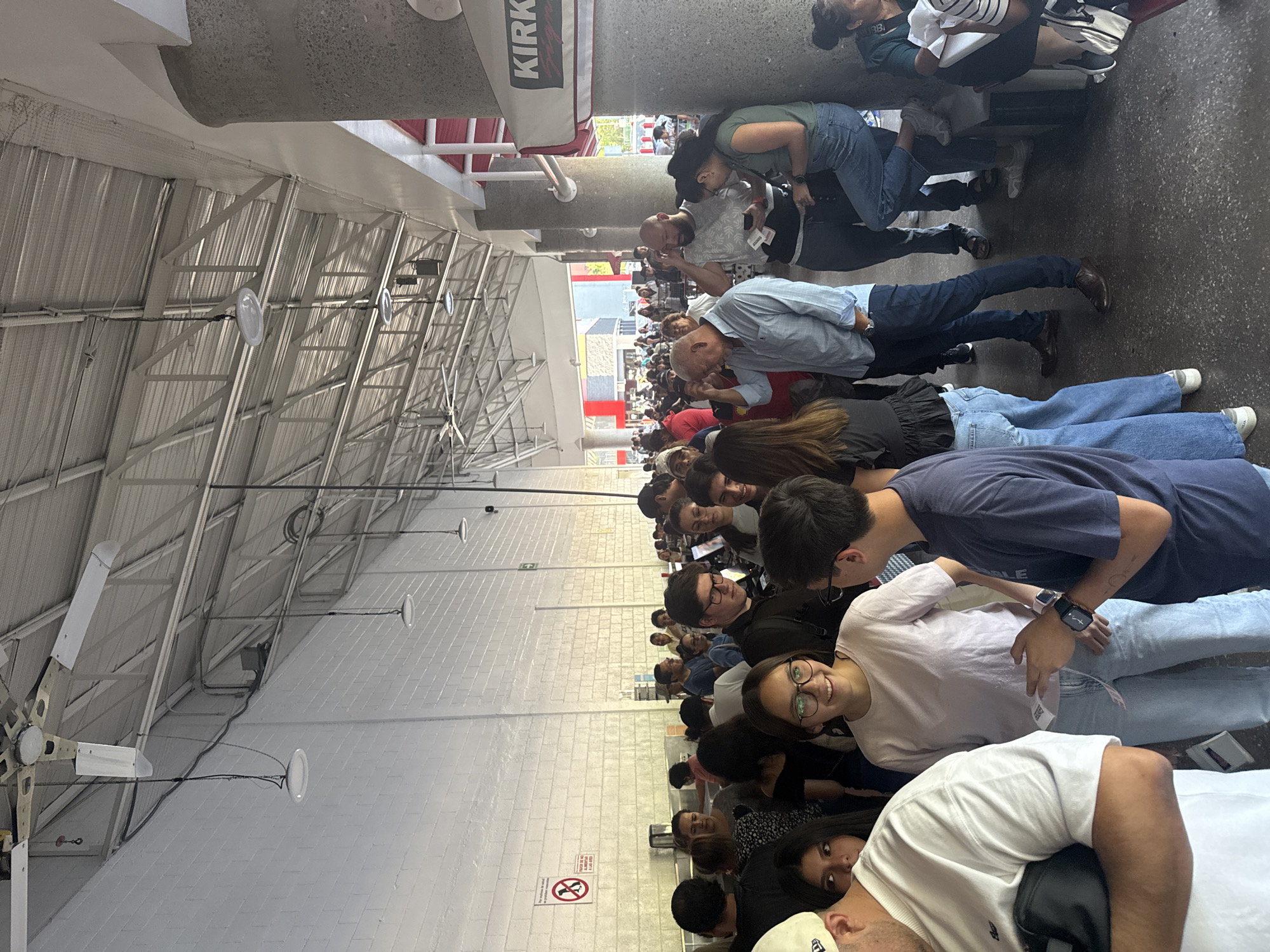 A photograph shows a long queue of people waiting in a Costco warehouse location in Mexico City, Mexico. The queue extends along a walkway alongside the entrance. The ceiling has industrial lighting and large ventilation fans. Several of the people are looking at the camera. The location is outdoors but covered.