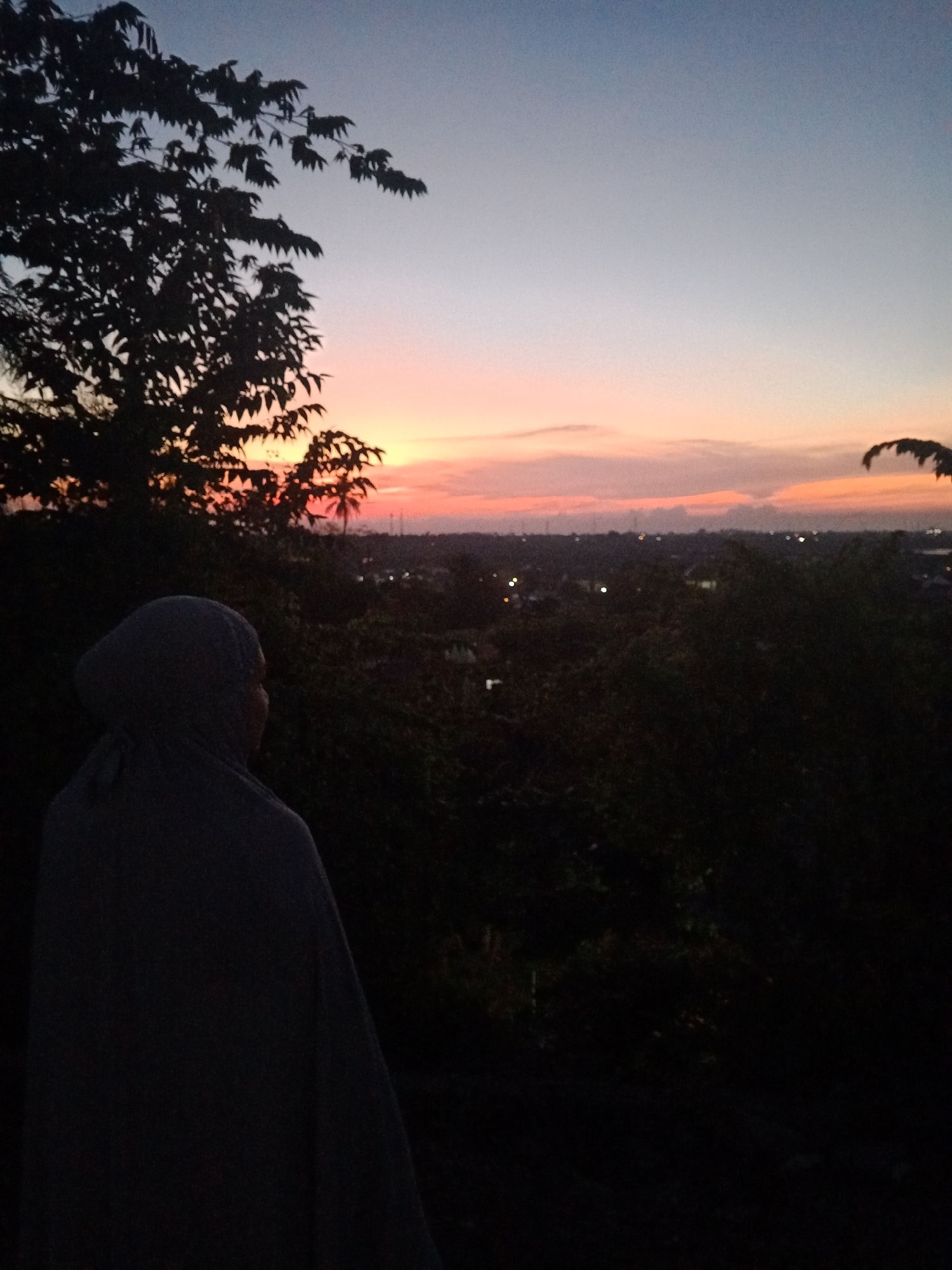 A woman in a headscarf stands in silhouette against a sunset sky over Lhokseumawe, Indonesia. The skyline has scattered buildings and a few streetlights are visible. In the foreground, there is a tree in the upper left corner and some vegetation that is blocking the view.