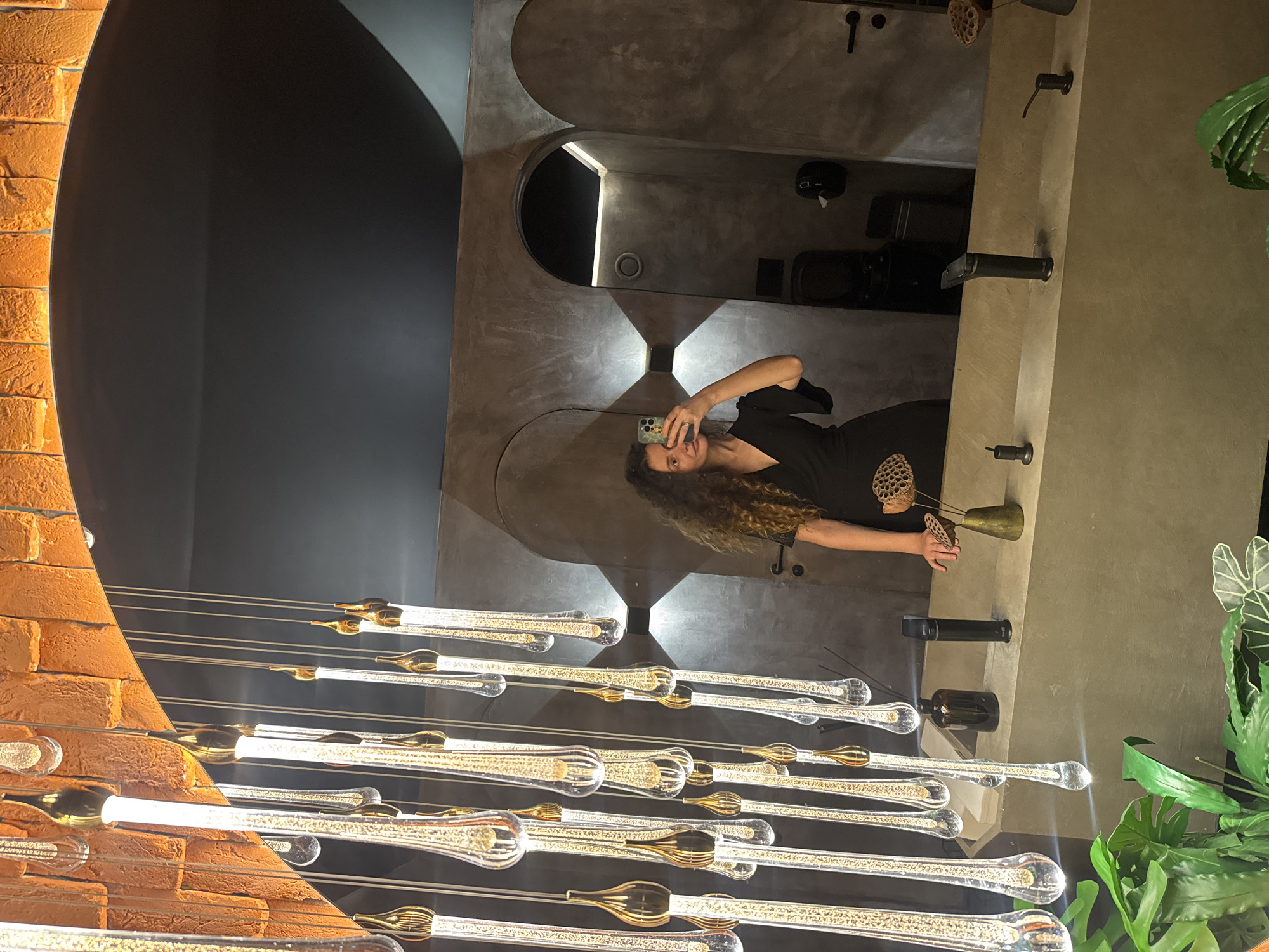 A woman with long, curly brown hair is taking a mirror selfie in a modern bathroom. She is wearing a black dress and is holding a smartphone in her right hand. The bathroom features a concrete sink and counter with black fixtures. A large mirror reflects the woman and the rest of the bathroom, which includes arched doorways and textured walls. Decorative pendant lights with glass droplets hang from the ceiling, and there are plants in the foreground. The lighting is dim and atmospheric.