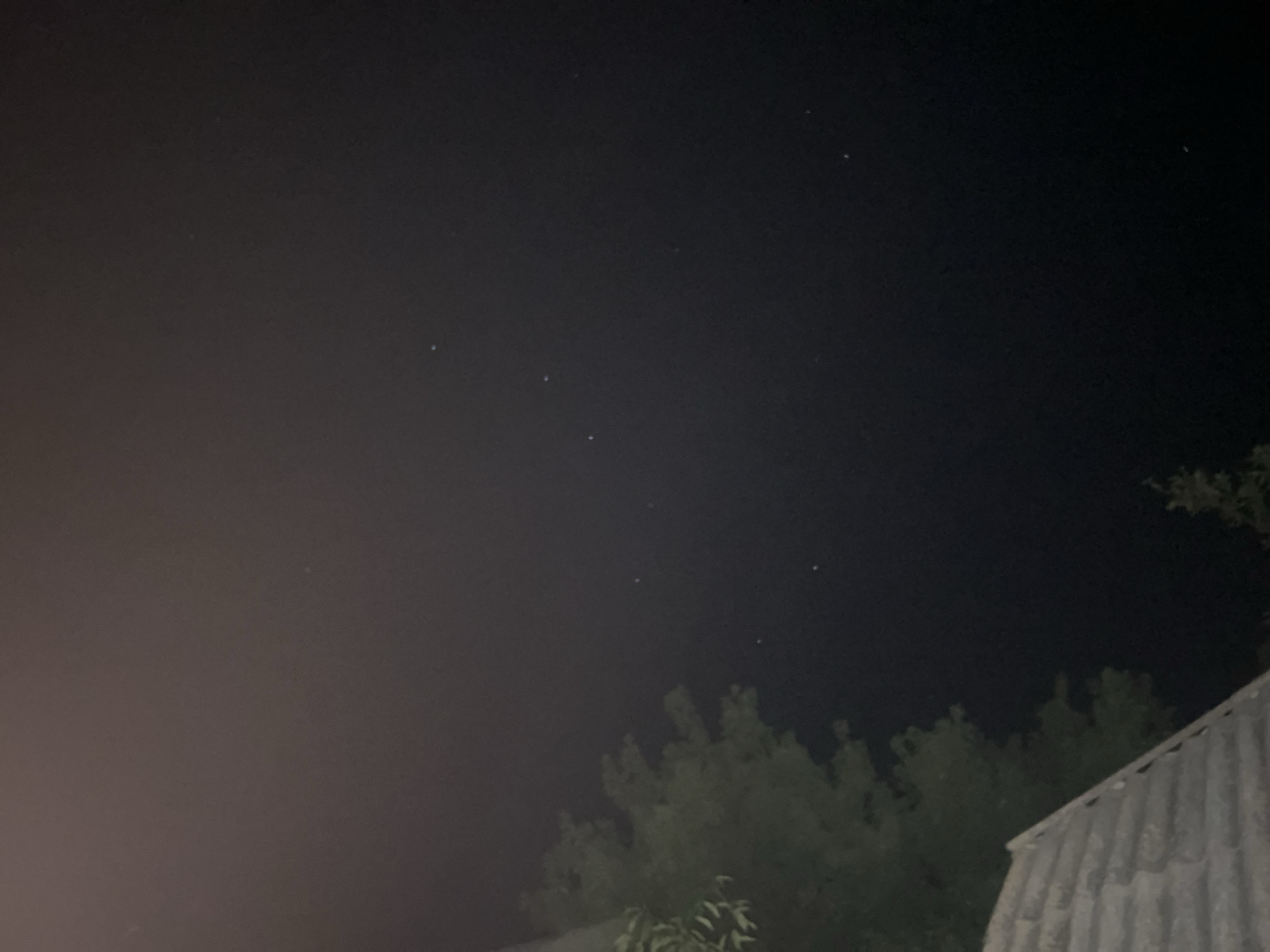 The image is a nighttime photograph taken outdoors, likely in the city of Zarichchia, Ukraine, judging by the context provided. The sky occupies most of the frame and is dark, with several small, bright points of light visible, which appear to be stars or possibly distant lights. The lower portion of the image shows the upper branches of trees, silhouetted against the sky and illuminated by a faint light source to the left. A portion of a corrugated roof structure is visible in the lower right corner, also dimly lit. No people or explicit actions are depicted. The overall scene suggests a rural or suburban setting at night, possibly with some light pollution from a nearby settlement.
