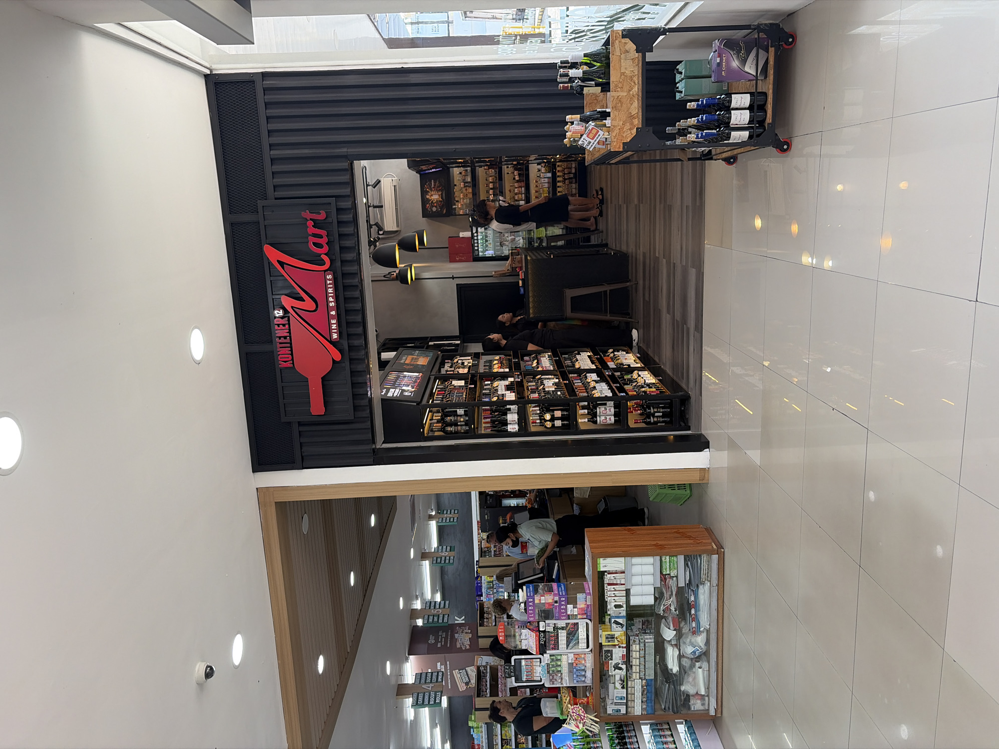 The image shows the entrance to a "KONTENER Mart WINE & SPIRITS" store, located in North Kuta, Indonesia. The store features a modern, industrial aesthetic with a corrugated metal facade and a large red "M" logo. Inside, aisles of wine bottles are visible on shelves and a display cart. Several employees and customers are present, with some appearing to be shopping or working at the counter. The floor is tiled and reflects the ambient lighting. The overall scene depicts a retail environment focused on alcoholic beverages.