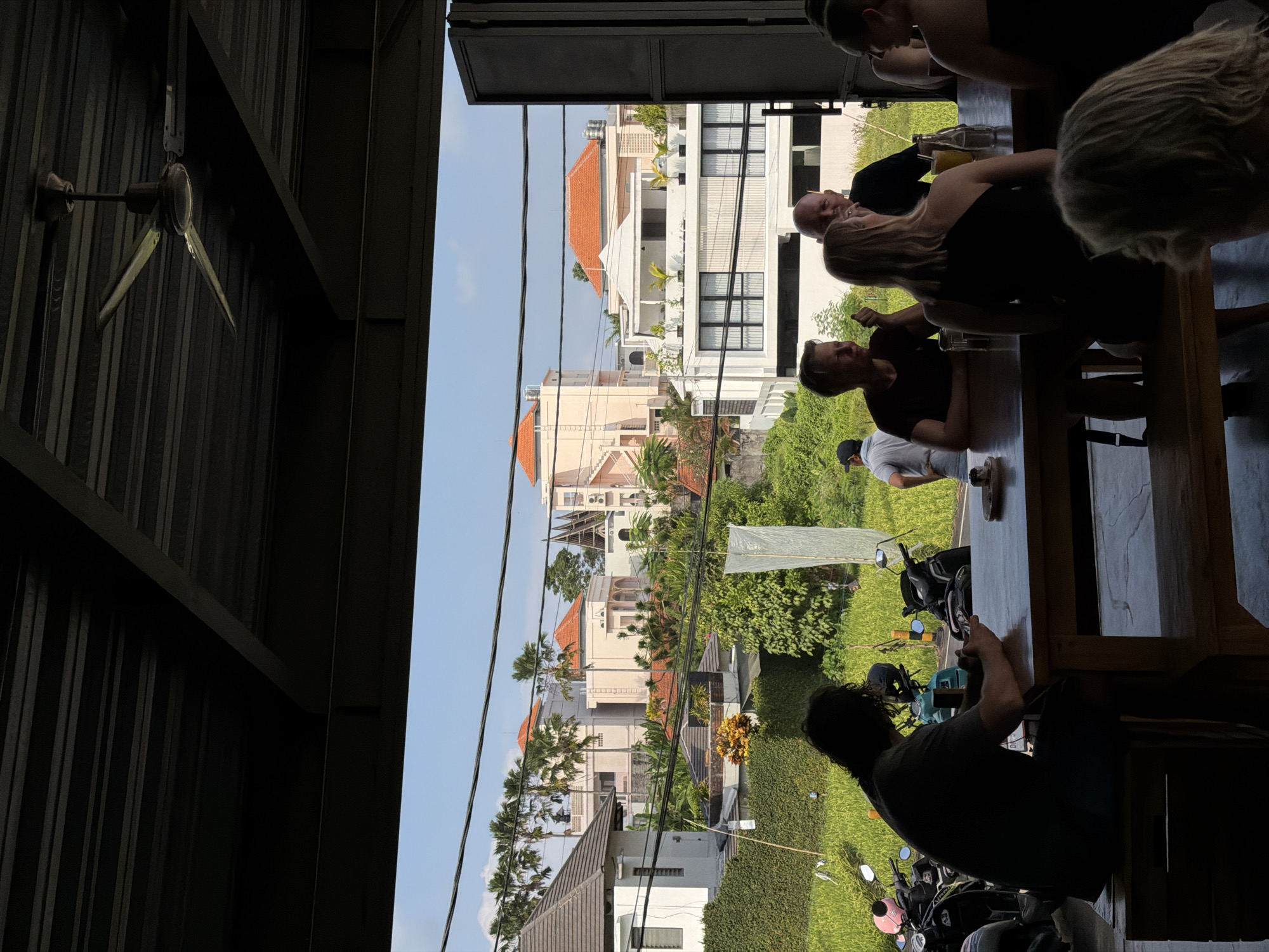 A group of individuals are seated at wooden tables, dining or conversing under a dark, corrugated metal roof. The foreground is dominated by the silhouettes of people and the tables, with a ceiling fan visible above. Beyond them, through an open-air structure, a vibrant scene unfolds. Modern villas with orange-tiled roofs and lush green landscaping are visible, interspersed with palm trees. A rice paddy in the middle ground adds to the rural, tropical atmosphere. In the background, a clear blue sky provides a backdrop to the scene. The overall impression is a blend of modern architecture and natural beauty, suggesting a relaxed, outdoor dining or gathering space in a tropical setting.
