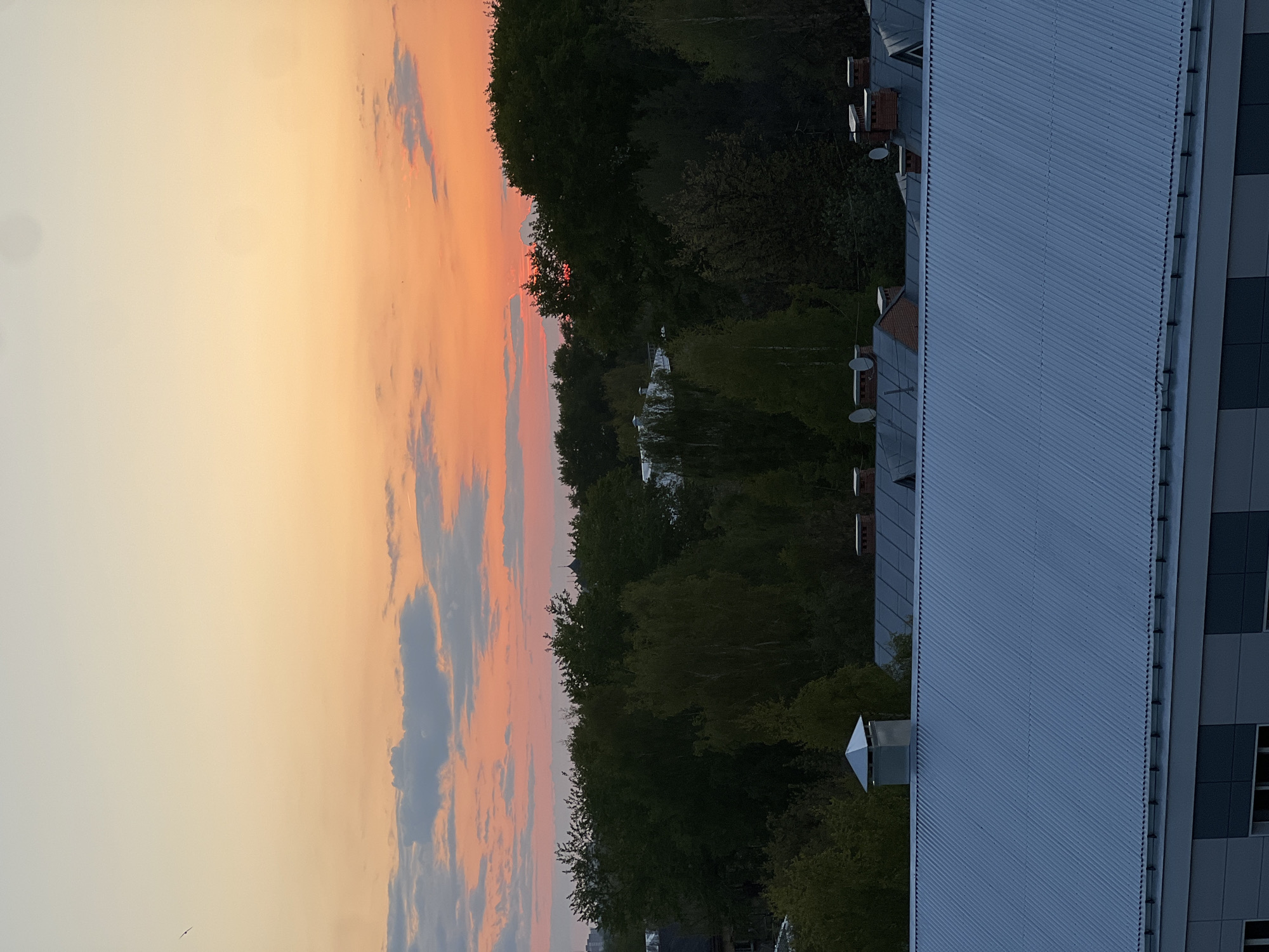 The image depicts a sunset in the city of Unknown, Unknown, taken from an elevated position, likely a building. A building with a corrugated metal roof dominates the right side of the frame. The skyline includes trees, and a body of water or open space is visible behind the trees. The sky displays a gradient of colors from pale yellow at the bottom to orange and pink toward the top. Scattered clouds are visible in the sky. The overall impression is of a sunset observed over an urban or suburban landscape.