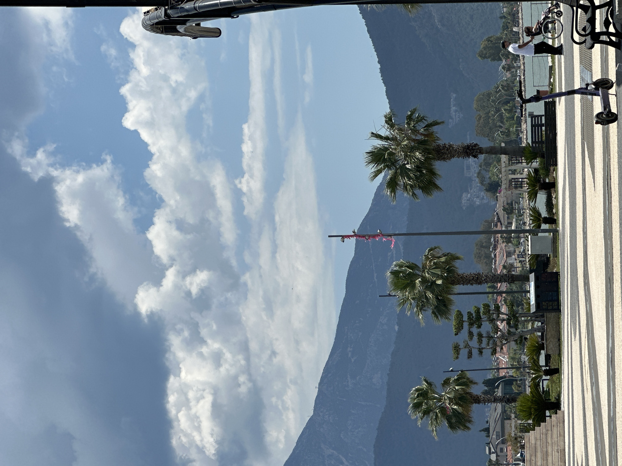 The scene is a view along a promenade in Fethiye, Turkey, on a bright day with a backdrop of mountains. The sky is mostly blue with intermittent white clouds. In the foreground, there is a paved walkway with a striped shadow pattern, lined with palm trees in planters. There are also benches and some electric scooters parked nearby. A man is standing near the edge of the promenade, seemingly photographing another person on a bicycle or in a wheelchair. The middle ground features the palm trees and glimpses of buildings. In the background, a large mountain dominates the view. Overhead, part of a structure is visible, possibly a street lamp or some sort of utility infrastructure.