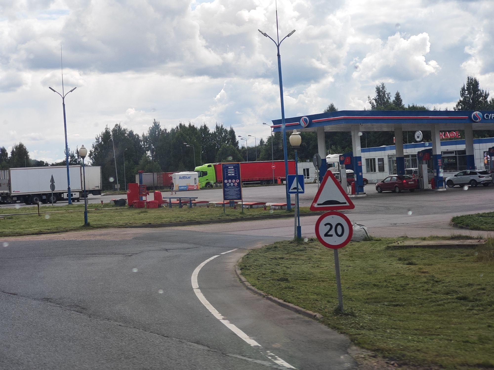 The image depicts a roadside scene near the city of Kresttsy, Russia. In the foreground, a paved road curves to the left, bordered by a grassy verge. A white line delineates the edge of the road. On the right, a speed limit sign indicates "20" within a red circle, and a triangular warning sign with a speed bump symbol is also visible.
In the background, a gas station is prominent, featuring a blue and red canopy over fuel pumps. Signs with Russian lettering, including "КАФЕ," are attached to the station building. Several vehicles are present, including a red car, a white SUV, and a white truck with a trailer. Further to the left, a green truck and another white truck with a trailer are parked. A lamppost with multiple lights stands near the center of the scene. Trees line the background, and the sky is filled with white, puffy clouds. The overall impression is a typical roadside service area.