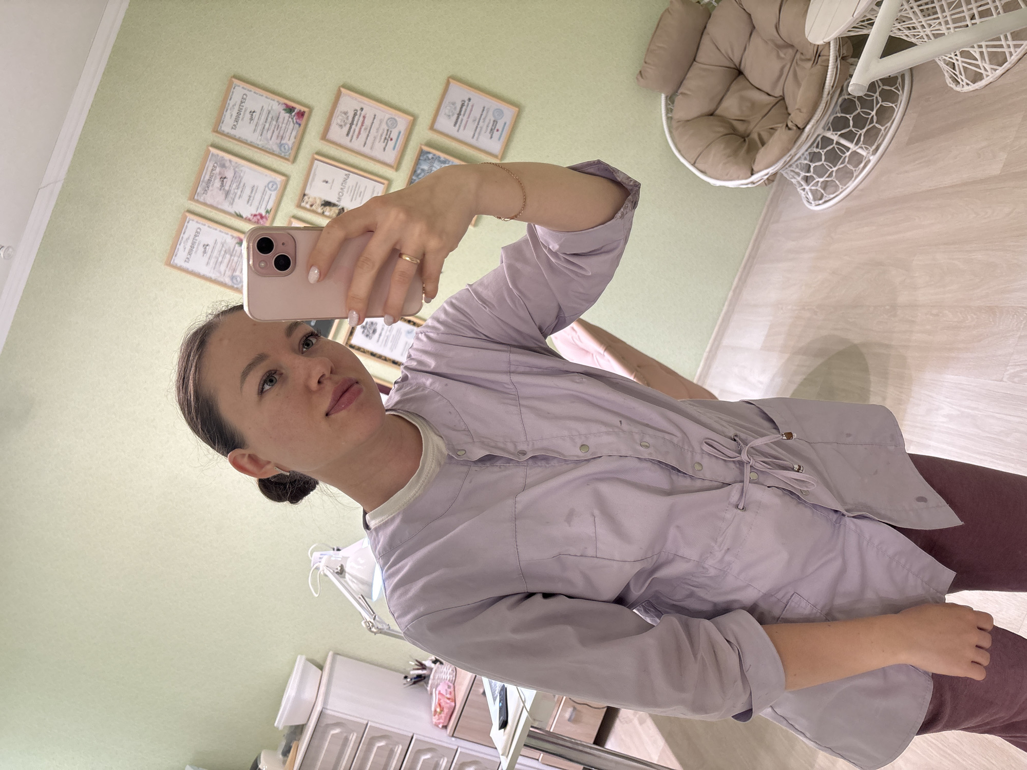 A woman in a light purple button-down shirt is taking a selfie with a pink smartphone. She has light skin, green eyes, and dark hair pulled back in a bun. She wears a bracelet on her left wrist and a ring on her left hand. The scene appears to be in a room within a business in Ферма, Russia, given the certificates on the wall and the medical examination table visible near the bottom of the frame. A white egg chair is visible in the upper right corner of the frame. There are cabinets along the wall in the lower left corner.