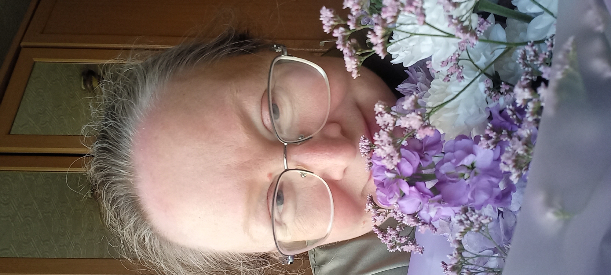 An adult woman with fair skin, graying hair, and glasses is looking at the camera. She is holding a bouquet of purple and white flowers close to her face. The background appears to be an indoor setting with wooden cabinets. The image is a close-up, portrait-style photograph. There is no discernible indication of the city of Izhevsk, Russia, in the image.