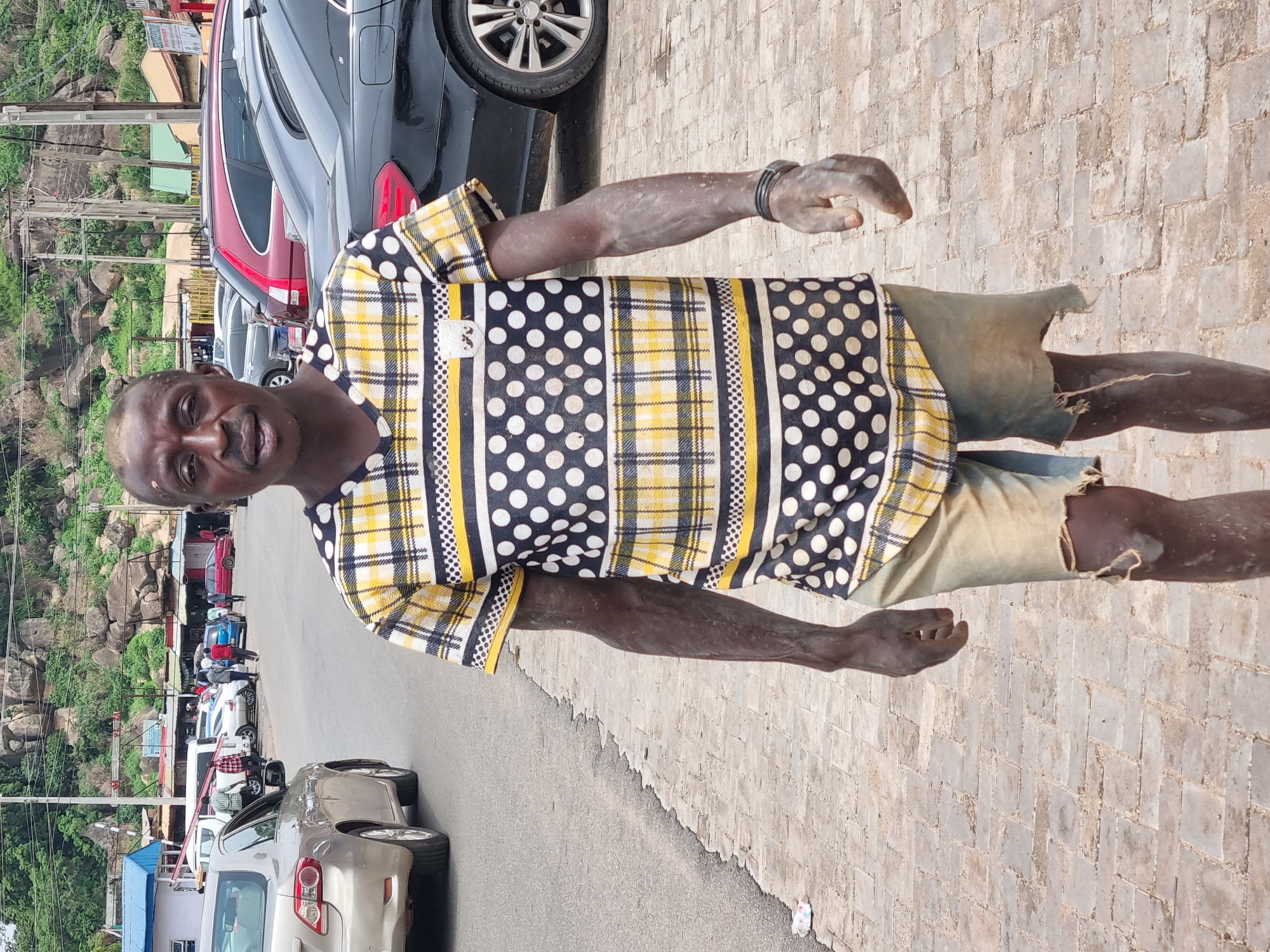 An outdoor, medium-shot, eye-level, full-body portrait captures a Black man walking towards the camera on a paved walkway. The man is wearing a black, white, and yellow plaid and polka-dot short-sleeved t-shirt with a small white pocket on the left chest. He also wears light-colored denim shorts that are ripped at the bottom. His arms and hands appear dusty.
The man stands in front of a street lined with parked cars and a rocky, vegetated hillside in the background. A variety of vehicles are visible, including a silver sedan on the left, a black sedan in the middle ground, and a maroon SUV behind it. In the distance, there are several small shops and stalls with awnings. The overall impression is of a street scene in a busy, perhaps informal, commercial area. The lighting suggests it is daytime, with some shadows indicating sunlight. The ground is paved with square interlocking bricks. The location is identified as Jos, Nigeria.