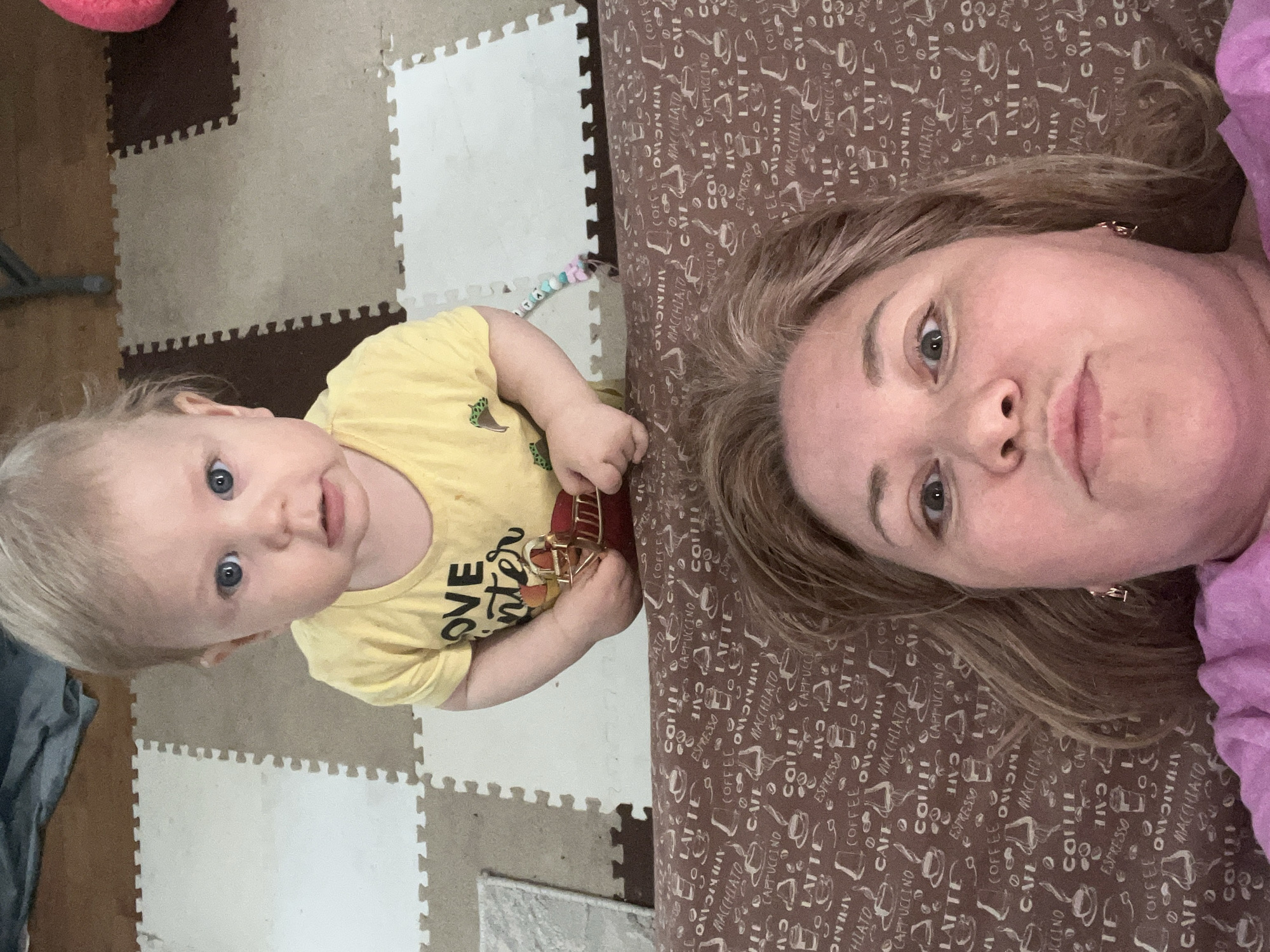 A woman and a young child are captured in a selfie-style photograph, potentially indoors, judging by the flooring and fabric backdrop. The flooring is composed of interlocking foam tiles, alternating between brown and beige/white. A portion of a dark-colored object is visible in the upper left corner. The child is positioned on the left side of the frame, wearing a yellow t-shirt with black text and holding a red object. The woman, on the right, is lying down. The fabric beneath her features a repeated pattern of coffee-related terms. The location, Vesyoloye, Russia, cannot be confirmed from the image.