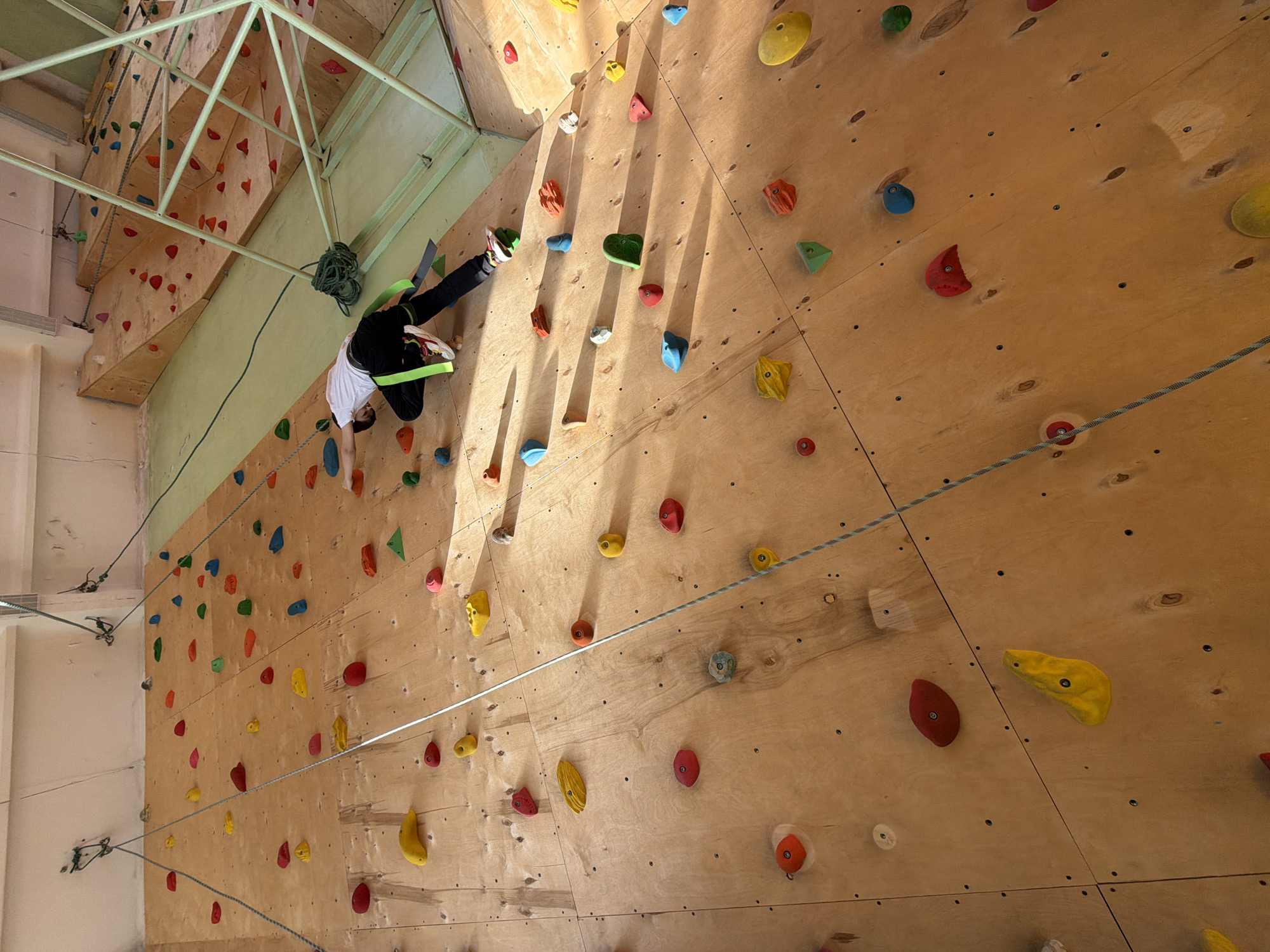 A person is climbing an indoor rock climbing wall. The climbing wall is constructed of wood and has a variety of colorful climbing holds attached. A safety harness and rope are visible, securing the climber. The climbing structure is situated in what appears to be a large indoor facility with metal support beams. The location is specified as the city of Izhevsk, Russia.