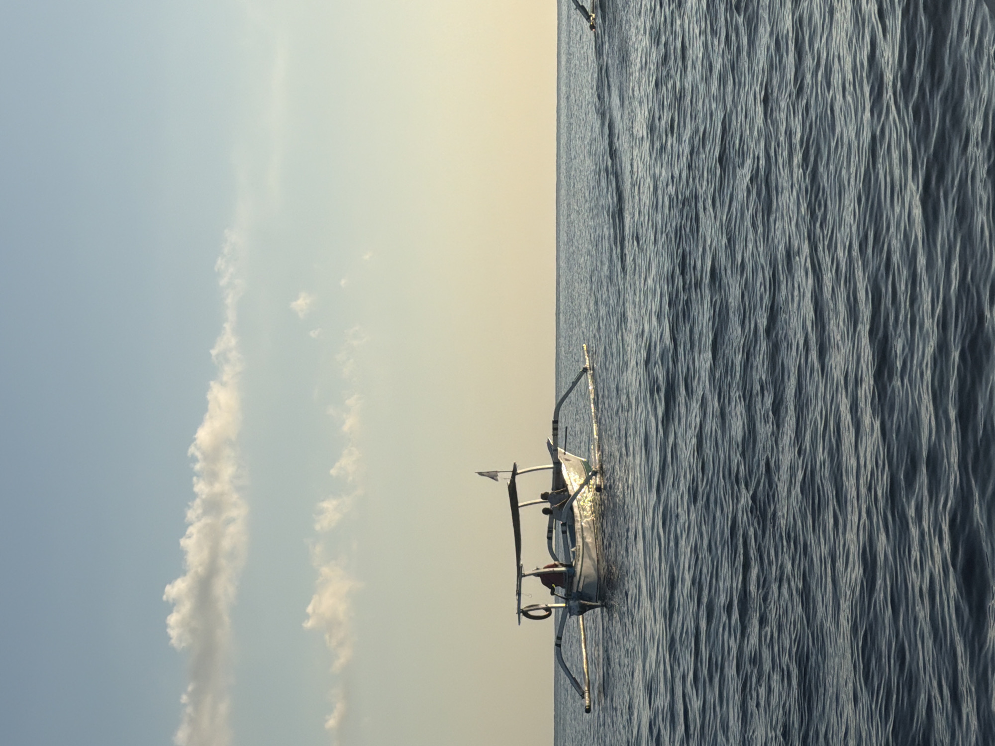 A traditional Balinese jukung boat with outriggers is on the ocean. Two people are visible in the boat, one appearing to steer. The water is dark blue with small waves. The sky is a pale blue with wispy white clouds and a soft gradient to a light yellow near the horizon. A very distant boat or object is visible on the right side of the frame. The image was taken in the waters off Seririt, Indonesia.