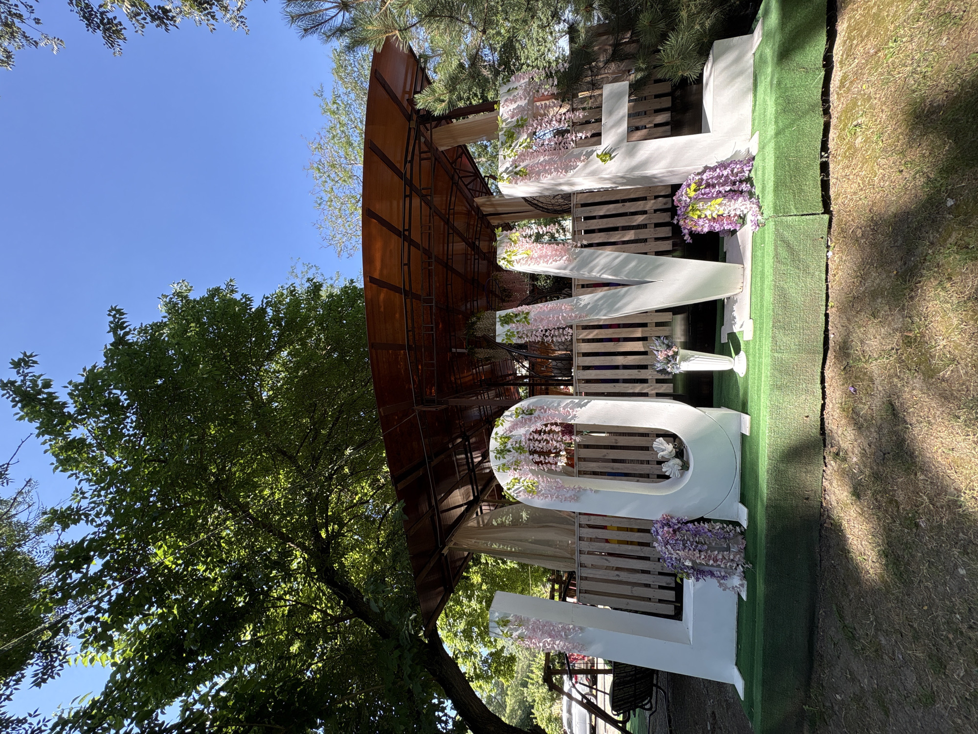 The image depicts a large, white, three-dimensional "LOVE" installation set outdoors, likely for a special event such as a wedding or celebration. The letters are adorned with artificial floral arrangements, primarily in shades of pink and purple, cascading down from the top and sides. The "O" and "V" are particularly decorated with these flowers.
The installation is placed on a raised platform covered in artificial green turf. Behind the "LOVE" letters, there is a wooden fence and a covered structure with a translucent, possibly orange or brown, roof supported by metal beams. Draped fabric, likely beige or cream-colored, hangs from the structure.
The surrounding environment is lush and green, with mature trees providing shade over the scene. The ground around the turf platform is covered in soil and grass. In the background, to the left, a portion of a playground structure with swings and a vehicle can be seen. To the right, pine trees frame the scene. The sky is clear and blue, indicating fair weather.
The overall impression is one of a decorated outdoor venue, possibly in a park or a garden area, designed to be visually appealing for photographs and events. The location is specified as the city of Novorossiysk, Russia.