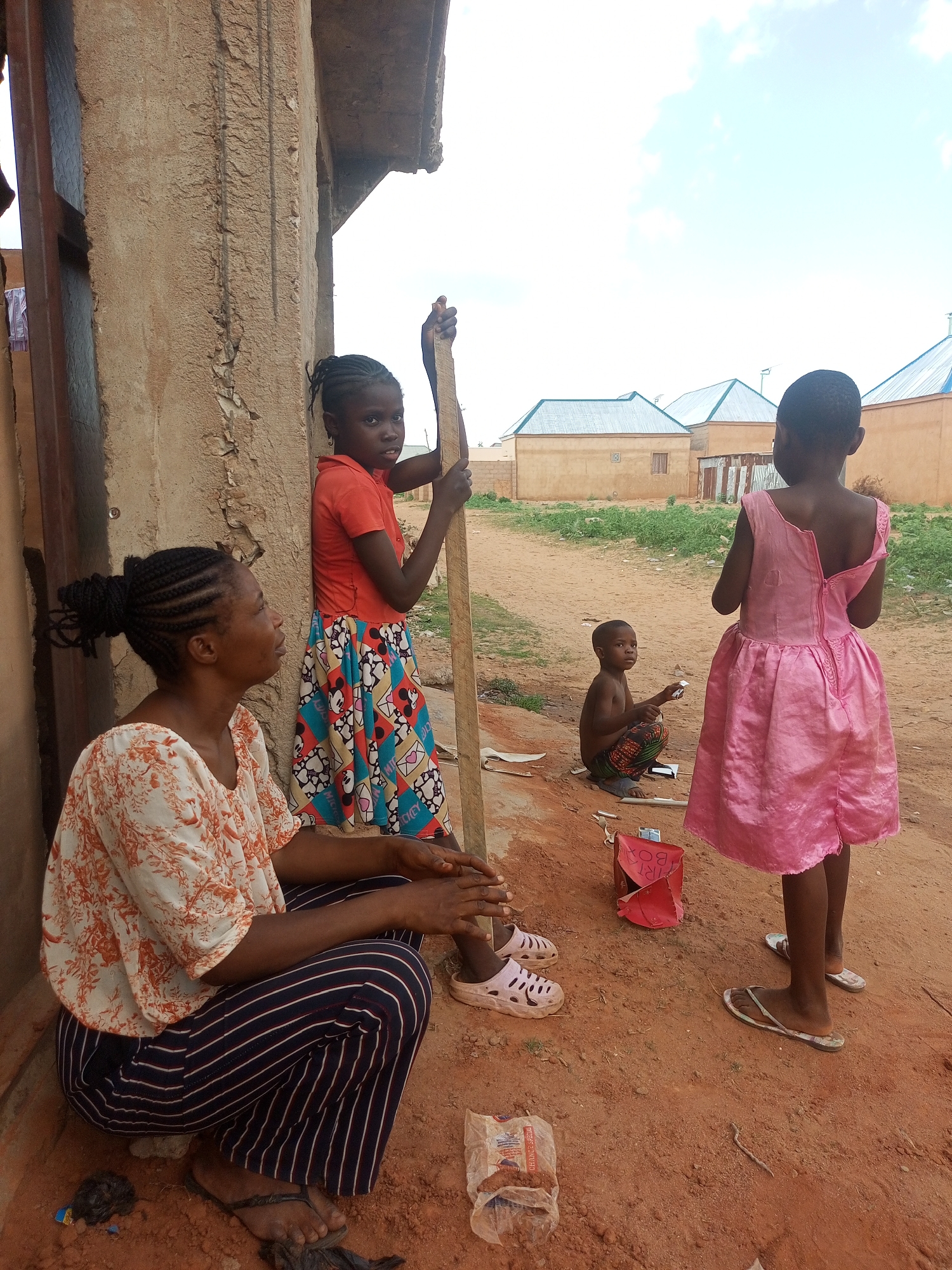 An outdoor scene in Tempure, Nigeria, features an adult woman and three children gathered on a dirt ground outside a weathered building wall.
On the left, an adult woman with braided hair is squatting, wearing a white top with an orange floral pattern and dark blue striped pants. She is looking to her right. Next to her, a young girl with braided hair stands, holding a long wooden stick. She wears an orange short-sleeved top and a colorful patterned skirt featuring cartoon characters like Mickey Mouse and hearts. She looks directly towards the viewer.
Further to the right, a small boy, shirtless, squats on the sandy ground, holding a small white object in his hands. Behind him and to his right, another young girl stands with her back to the viewer, wearing a bright pink sleeveless dress with a visible zipper running down the back. She appears to be looking towards the background.
The ground is predominantly reddish-brown dirt. Various small items are scattered on the ground, including pieces of paper, a red crumpled box with "IRT BOX" visible, and a clear plastic bag with "GHANA SPECIAL" printed on it. Footwear includes flip-flops on the woman and the girl in pink, and white sandals on the girl in the patterned skirt.
In the background, under a bright, cloudy sky, several single-story houses with light-colored walls and shiny blue or green corrugated metal roofs are visible, indicating a residential area. Sparse green vegetation grows on the dirt path between the foreground and the houses. The time of day appears to be daytime, with clear, bright conditions.