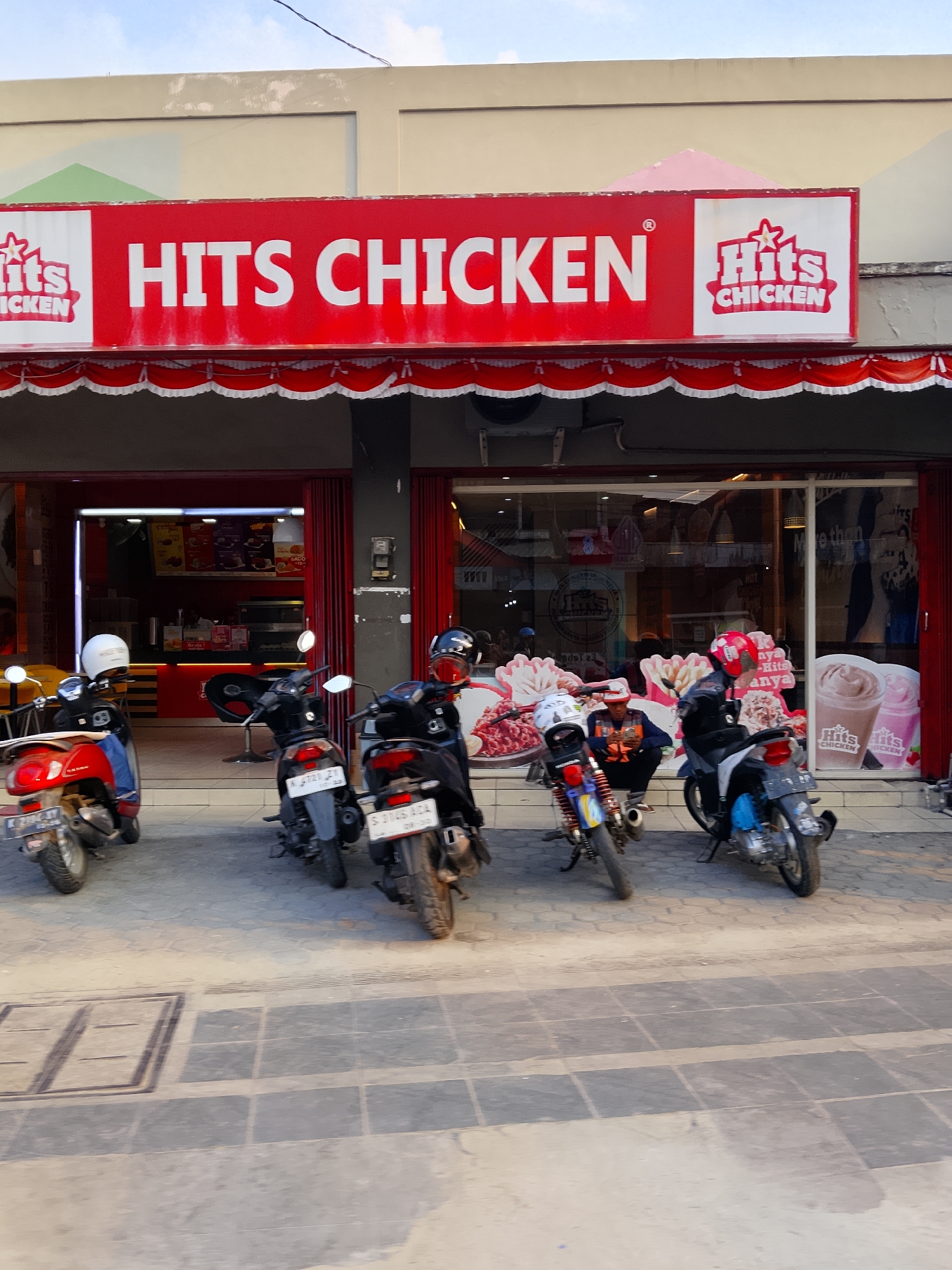 The image shows the storefront of a "Hits Chicken" restaurant in Balun, Indonesia. A red and white awning with a ruffled edge stretches across the top of the entrance, and the restaurant's name is prominently displayed in large white letters on a red background. Two logos featuring "Hits Chicken" are visible on either side of the main signage. In front of the restaurant, several motorcycles are parked on the paved walkway. Two men are visible inside the restaurant, one of whom appears to be a staff member at a counter. Large posters advertising food and drinks are displayed on the window and inside the establishment. The overall scene depicts a typical street view of a commercial establishment in Indonesia.