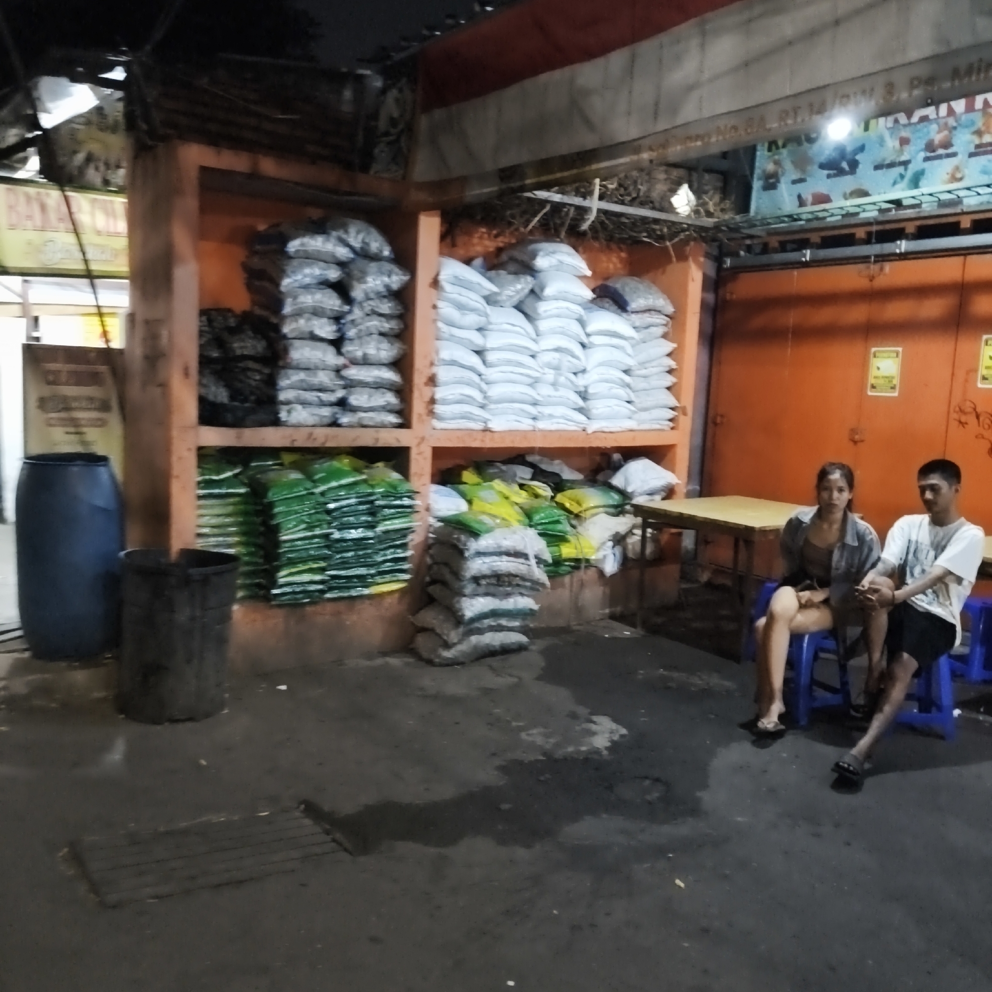 In South Jakarta, Indonesia, at night, a dimly lit scene depicts an outdoor vendor stall. Stacks of bagged goods, some appearing to be charcoal or similar materials, are arranged on shelves. On the ground, large plastic barrels are visible, along with smaller trash cans. Two young adults, a woman and a man, are seated on blue plastic stools, facing the viewer. The woman wears a grey jacket over a dark top and shorts, with sandals. The man is in a white t-shirt and black shorts, also wearing sandals. They appear to be waiting or taking a break. The environment suggests a street-side or open-air market setting, with a shuttered storefront in the background.