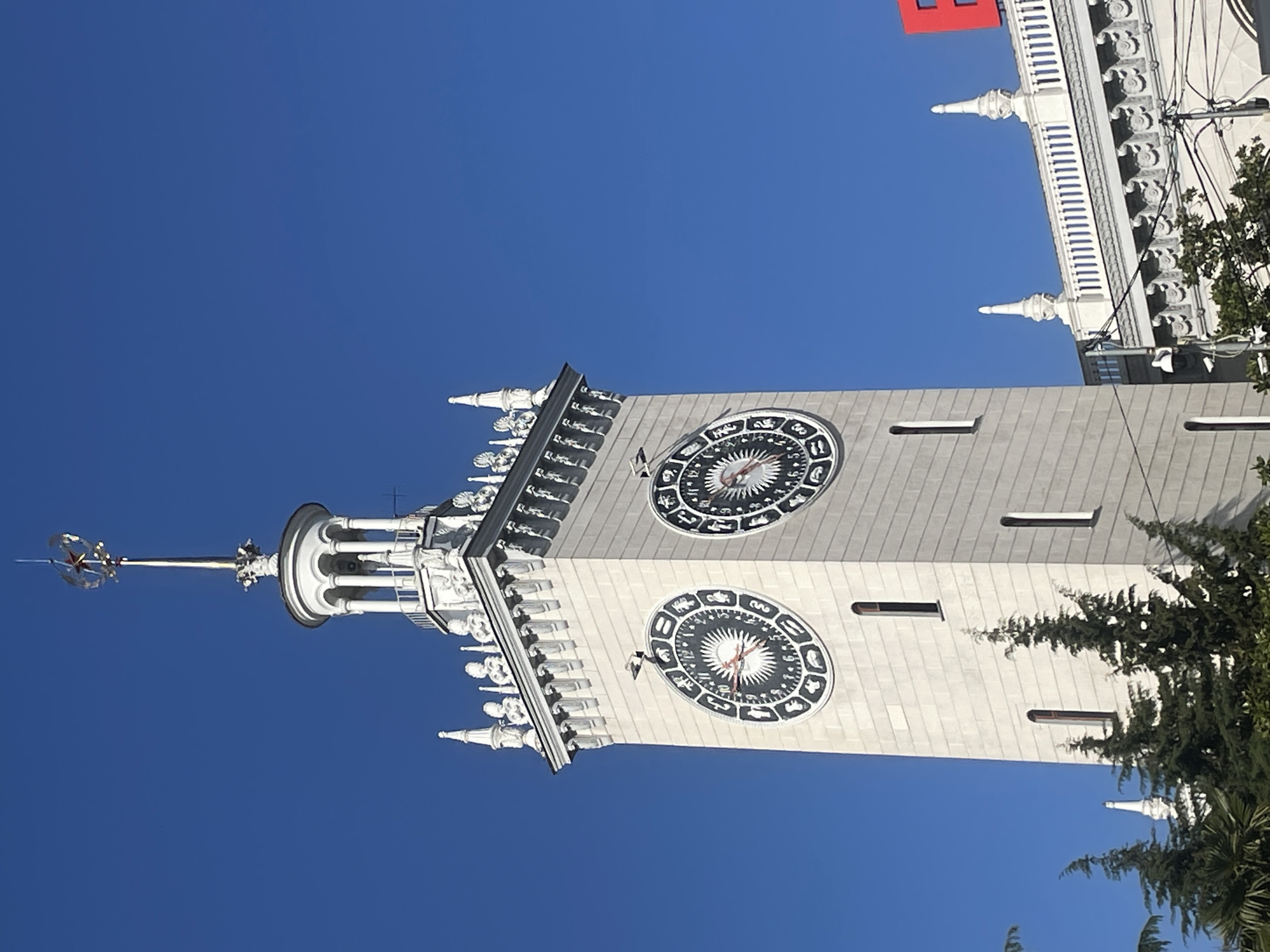 The image depicts a low-angle view of the Sochi Marine Station clock tower against a clear blue sky. The tower is white or light gray and features two large clock faces with zodiac symbols. Architectural details include ornate carvings, spires, and a star-topped spire at the top. A red flag is visible atop an adjacent building. Some green foliage is visible on the right side of the frame. The location is the city of Sochi, Russia.