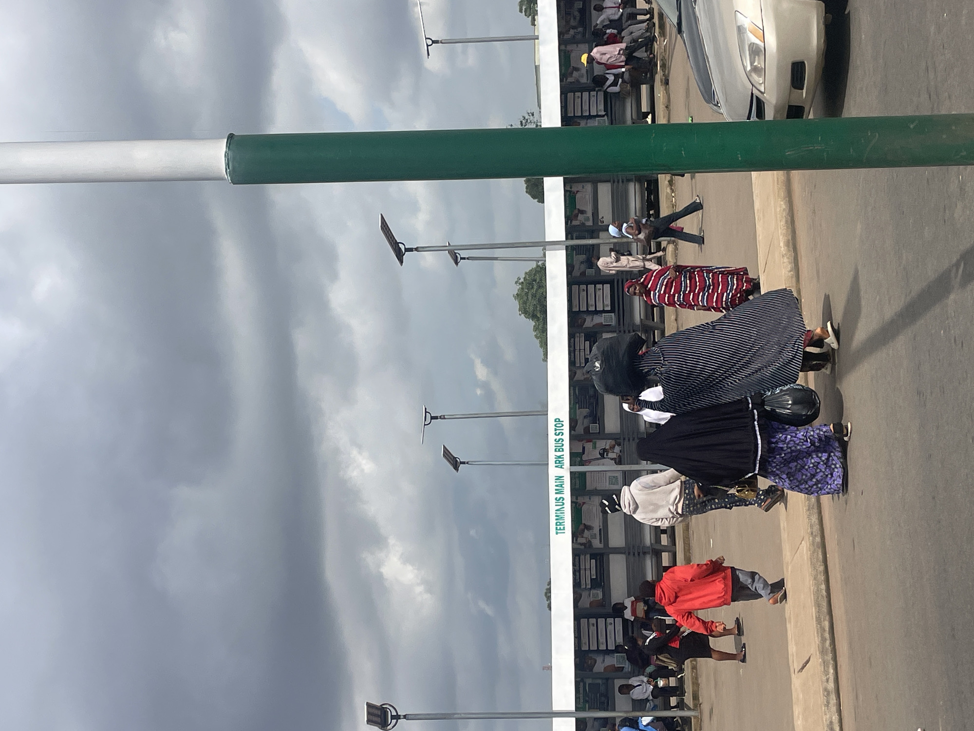 The image depicts a bus stop scene in Jos, Nigeria, under a cloudy sky, suggesting it might be morning or late afternoon. The primary subjects are a group of people, likely waiting for or disembarking from a bus. They are dressed in various outfits, including traditional African attire and casual wear, indicating a mixed demographic.
Objects visible include a silver car parked in the background, a bus stop shelter with multiple seating areas and what appear to be display boards, and streetlights with solar panels. A prominent feature is a tall, green and white striped pole that bisects the image, possibly a signpost or a barrier.
The activity appears to be people congregating at a transportation hub. Some individuals are standing, while others are seated within the shelter. The general atmosphere seems calm, with no overt signs of distress or urgency. The cloudy weather contributes to a muted, diffused lighting condition.
Notable details include the text "TERMINUS MAIN ARK BUS STOP" printed vertically on the bus stop shelter, confirming the location. The presence of people dressed in a variety of clothing styles suggests a typical day in a Nigerian city. The overall scene captures a moment of everyday life at a public transport facility.