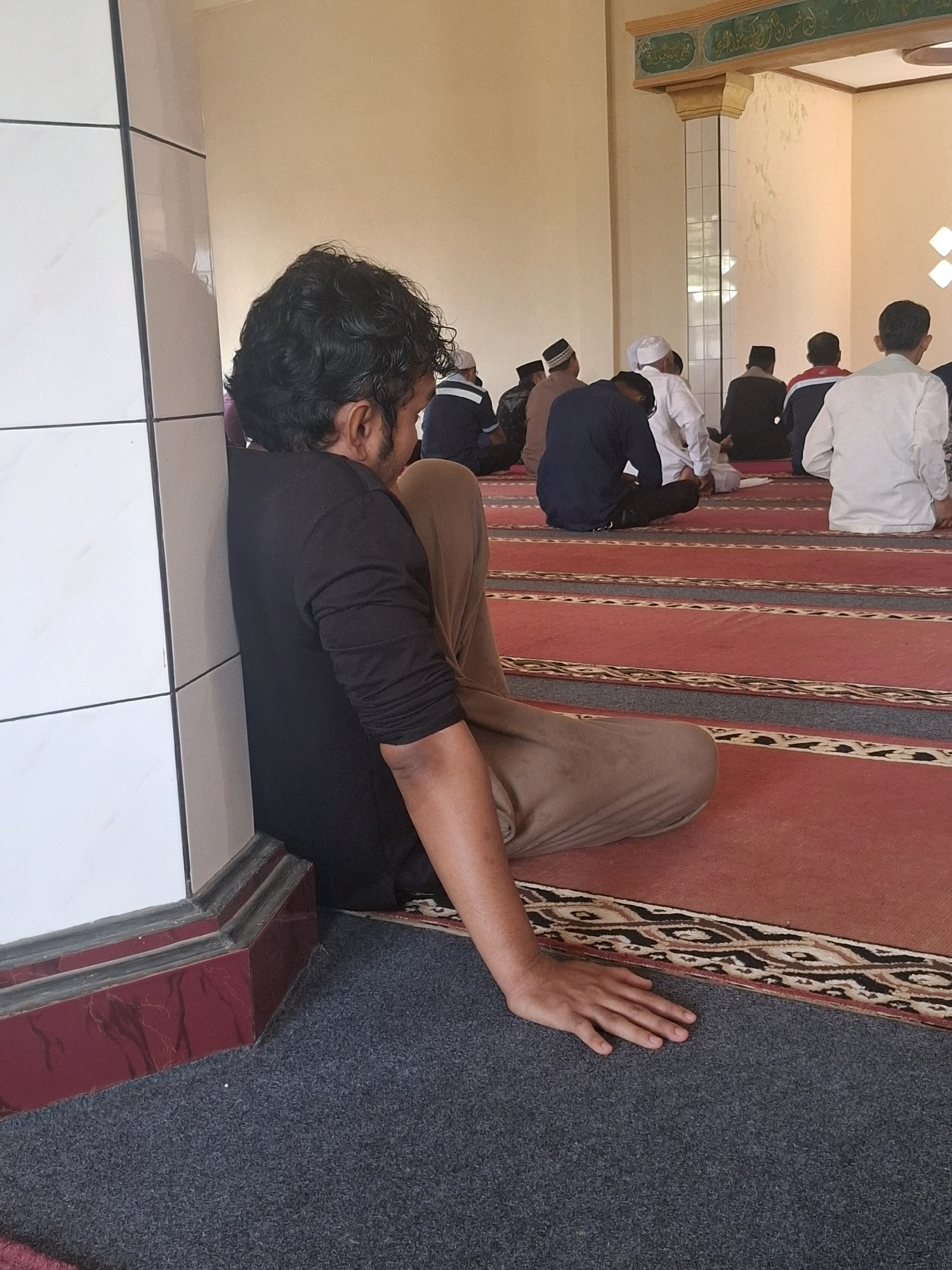 In Ciheulang Tonggoh, Indonesia, a group of men are seated on the floor inside a mosque. The man in the foreground, leaning against a white tiled pillar, wears a black long-sleeved shirt and light brown pants. His arm rests on the floor beside him. The other men are arranged in rows on a red carpet with patterned borders, facing the front of the mosque. They are dressed in various outfits, including white shirts, black pants, and skullcaps. The mosque interior features a decorative ceiling with inscriptions.
