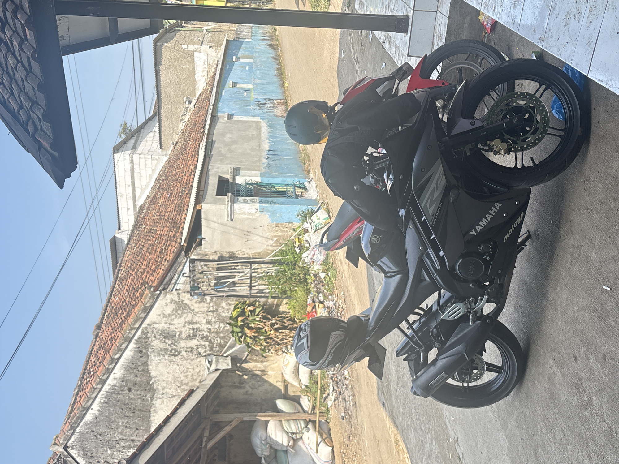 A black Yamaha motorcycle is parked on a paved surface in front of a weathered building with a blue facade and a red-tiled roof. A red motorcycle is partially visible behind the black one. A black helmet with a white and grey design rests on the rear seat of the black motorcycle. The handlebars of the black motorcycle are draped with a black jacket. In the background, a dirt road is visible, lined with more buildings and piles of sacks. Overhead power lines are strung across the clear blue sky. The scene appears to be an outdoor area in a village setting, possibly in Pasirlangu, Indonesia, given the architectural style and the presence of motorcycles which are common modes of transportation.