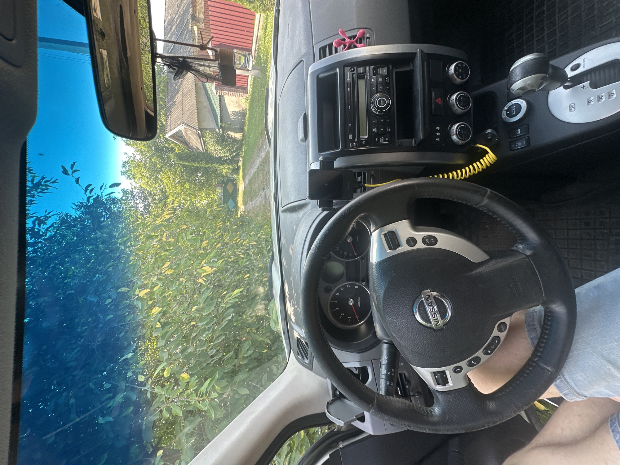 The image shows the interior of a Nissan X-Trail vehicle, viewed from the driver's perspective, parked on a dirt road in Shatava, Ukraine. The dashboard, steering wheel, and center console are clearly visible. Outside the car, lush green foliage and trees line the road, with houses visible in the background. The windshield displays a reflection of the sky and trees, indicating it is daytime. A person's legs, clad in denim shorts, are visible in the lower left corner, suggesting they are the driver or a passenger. A yellow coiled cable is attached to a phone mount on the dashboard.