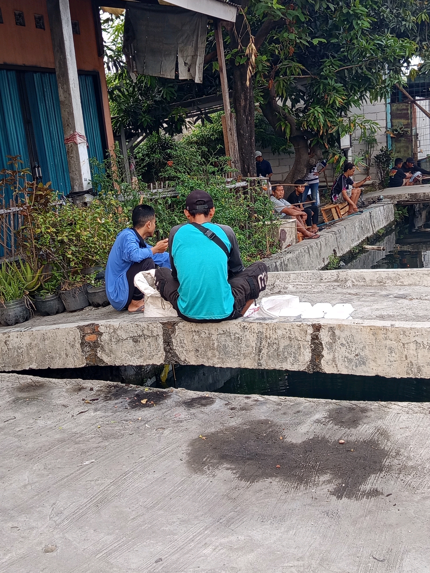 In Karawang, Indonesia, two men are seen sitting on a concrete ledge overlooking a narrow canal. One man, wearing a blue long-sleeved shirt and dark pants, is seated with his legs tucked underneath him. The other man, facing away from the camera, wears a turquoise t-shirt and black shorts. In front of them on the ledge are several white plastic containers and a white plastic bag. The canal below is dark and appears to contain some debris. Behind them, a building with a blue corrugated metal door is visible, along with lush green foliage and trees. Further in the background, other individuals are seen sitting by the canal, some with fishing rods, suggesting a communal fishing spot. The overall scene depicts a casual outdoor gathering by the water.