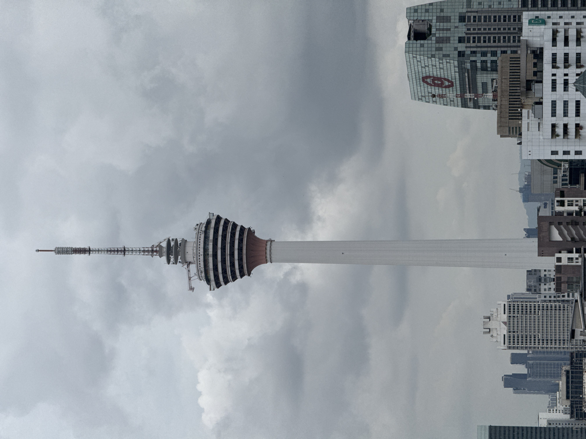 The image shows the Kuala Lumpur Tower (Menara Kuala Lumpur), a communications tower located in Kuala Lumpur, Malaysia, from an elevated position. The tower is the central focus, standing tall against a cloudy sky. In the background, there are several high-rise buildings, typical of a modern city skyline. The architecture of the buildings varies, and there appears to be a mix of residential and commercial structures. The lighting suggests an overcast day, and the image is shot in a way that captures both the grandeur of the tower and its urban setting.